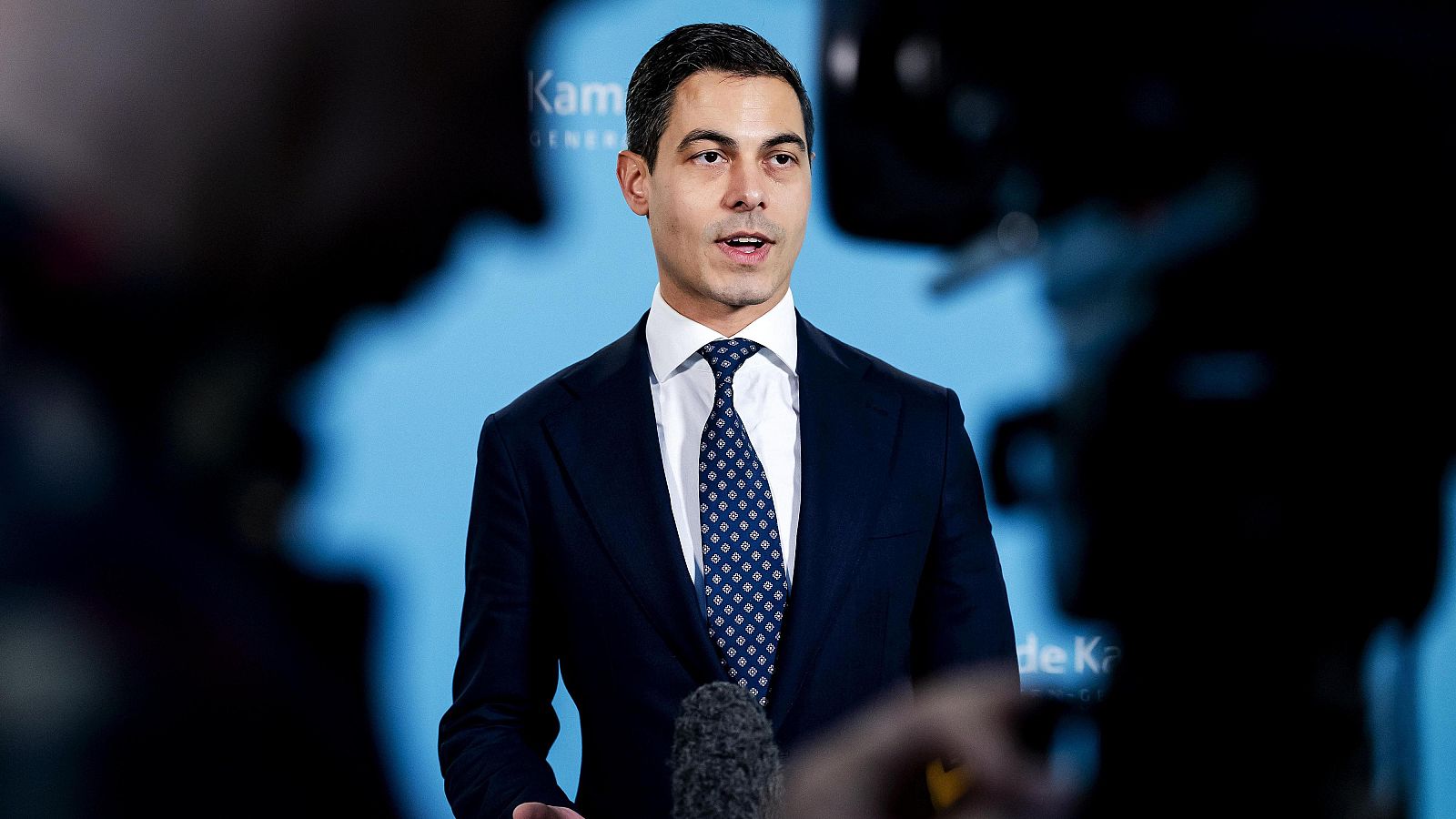 Rob Jetten, con traje azul y corbata estampada, habla ante micrófonos con un fondo azul y un mapa estilizado. La imagen muestra al político holandés en un evento.