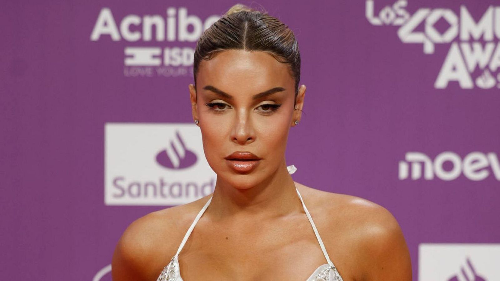 La actriz Jedet, con un moño alto y maquillaje definido, posa en la alfombra con un vestido blanco y detalles de encaje, frente a un fondo con logotipos.