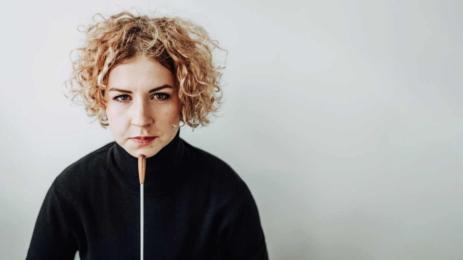 Barbara Dragan, con el pelo rubio rizado y un jersey negro, sostiene una batuta frente a su barbilla mientras mira directamente a la cámara, con un fondo blanco.