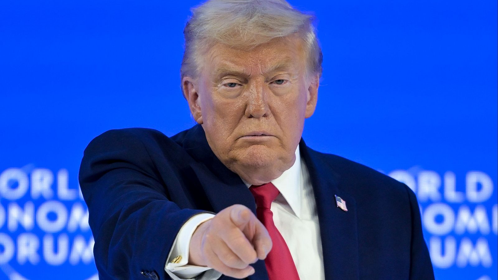 Donald Trump, con traje oscuro y corbata roja, señala a la cámara con el dedo índice, mostrando una expresión seria frente a un fondo azul con la inscripción "WORLD ECONOMIC FORUM".