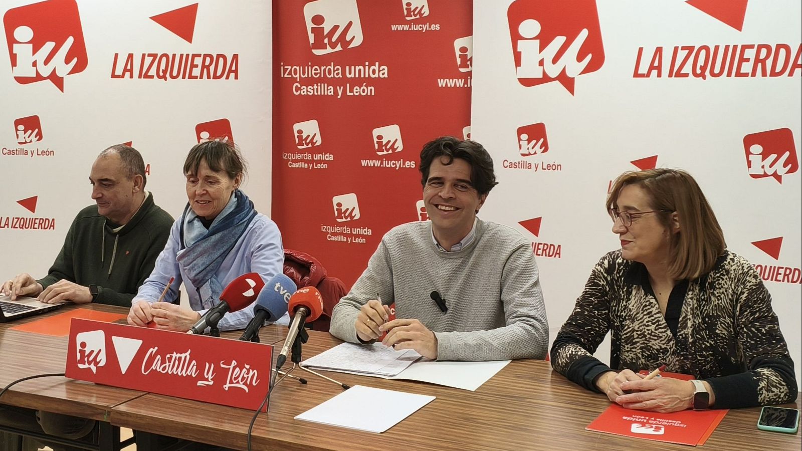 En una mesa, un hombre con jersey gris claro y otros tres individuos participan en un evento. Se observan micrófonos, papeles y un cartel con el nombre de una organización política.