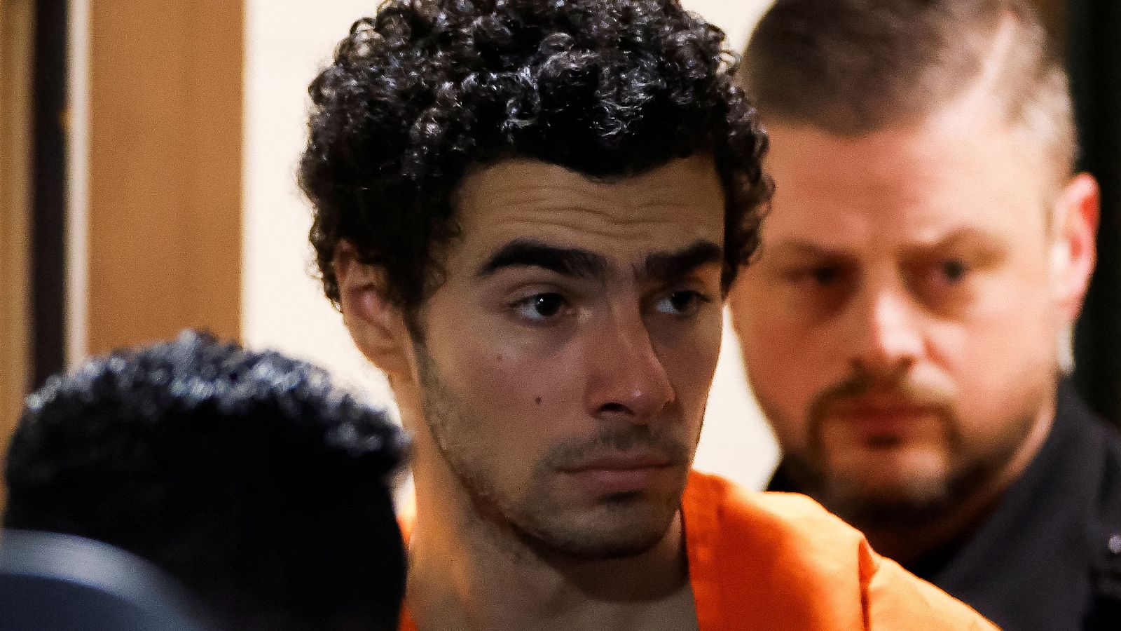 Hombre con mono naranja y cabello oscuro, expresión seria, mira hacia abajo. Fondo desenfocado con figura en ropa oscura, posible agente de seguridad. Entorno institucional.