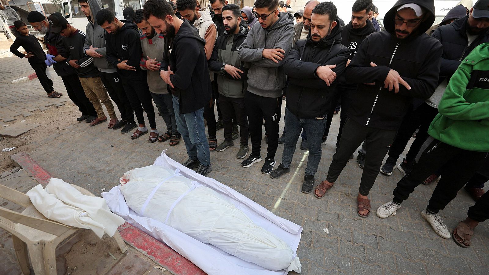 Hombres vestidos con ropa informal rodean un cuerpo cubierto con una tela blanca, mostrando respeto y tristeza en un espacio al aire libre, posiblemente durante un funeral.