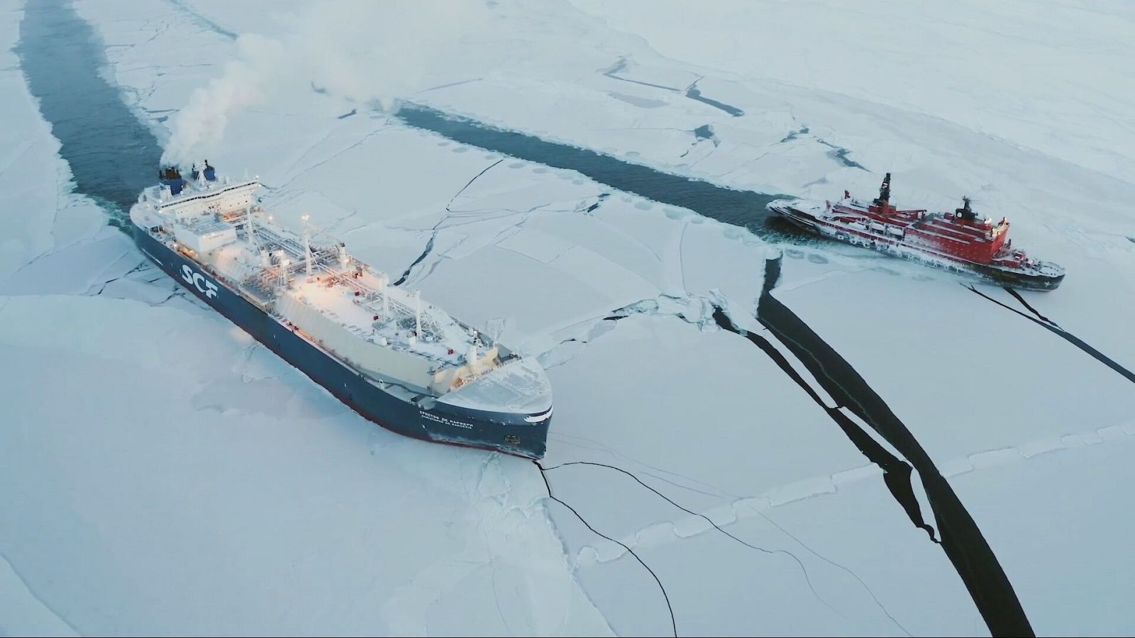En un entorno ártico helado, un buque de carga y un rompehielos navegan entre el hielo fracturado, con el cielo cubierto y sugiriendo condiciones climáticas frías.