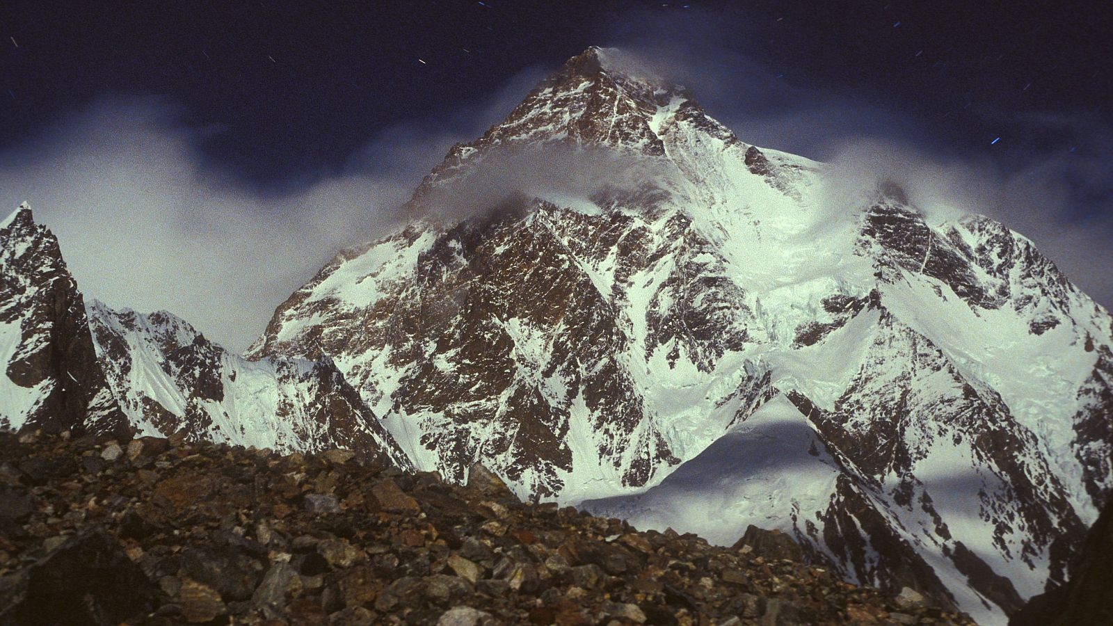 Una fotografía muestra la cara sur del K2, una montaña imponente cubierta de nieve, con zonas rocosas visibles y nubes que rodean la cima, difuminando su contorno contra el cielo.