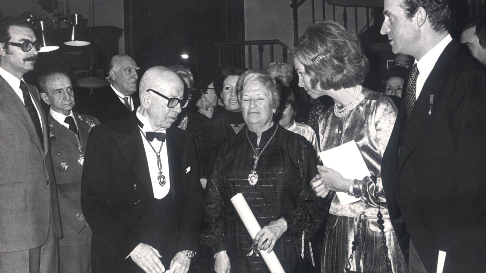 Imagen en blanco y negro de Carmen Conde tras su discurso, acompañada por la Reina Sofía y el Rey Juan Carlos I, en un evento formal. Se observan otros asistentes, todos elegantemente vestidos.