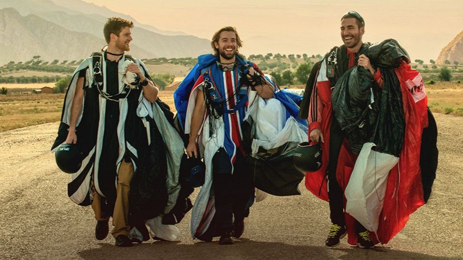 Tres paracaidistas, sonrientes y con cascos, posan en un camino montañoso. Uno de ellos viste un traje con los colores de una bandera, mientras que los otros dos llevan trajes de colores contrastantes.