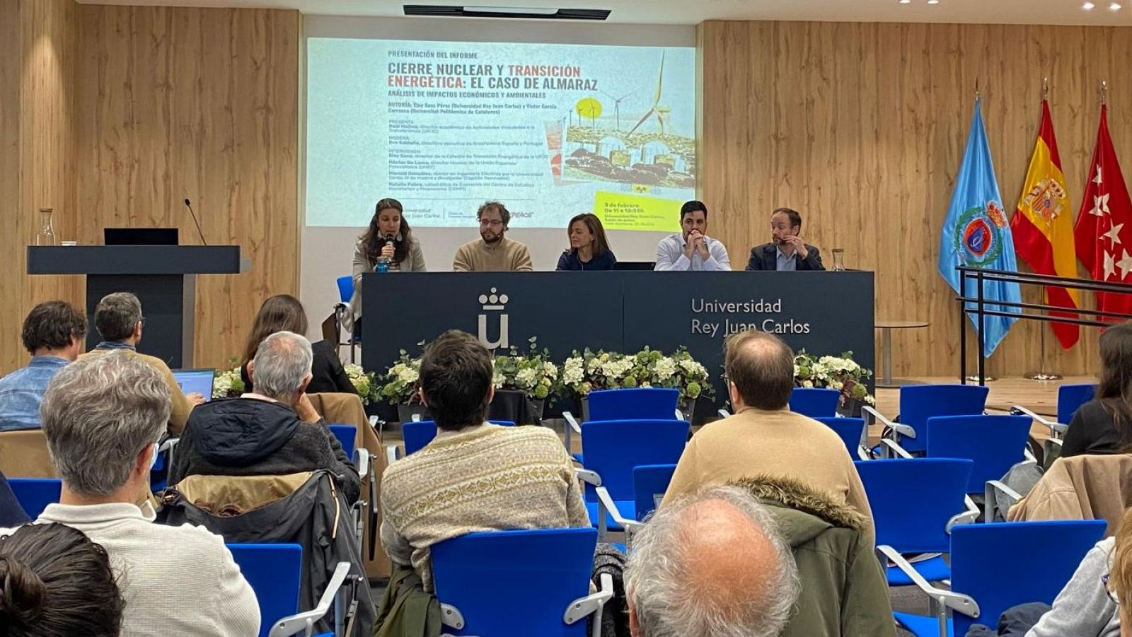 Un panel de expertos presenta un informe sobre el cierre nuclear y la transición energética en un evento académico. Se observa una mesa con micrófonos, flores y el logo de la universidad, así como una audiencia atenta.