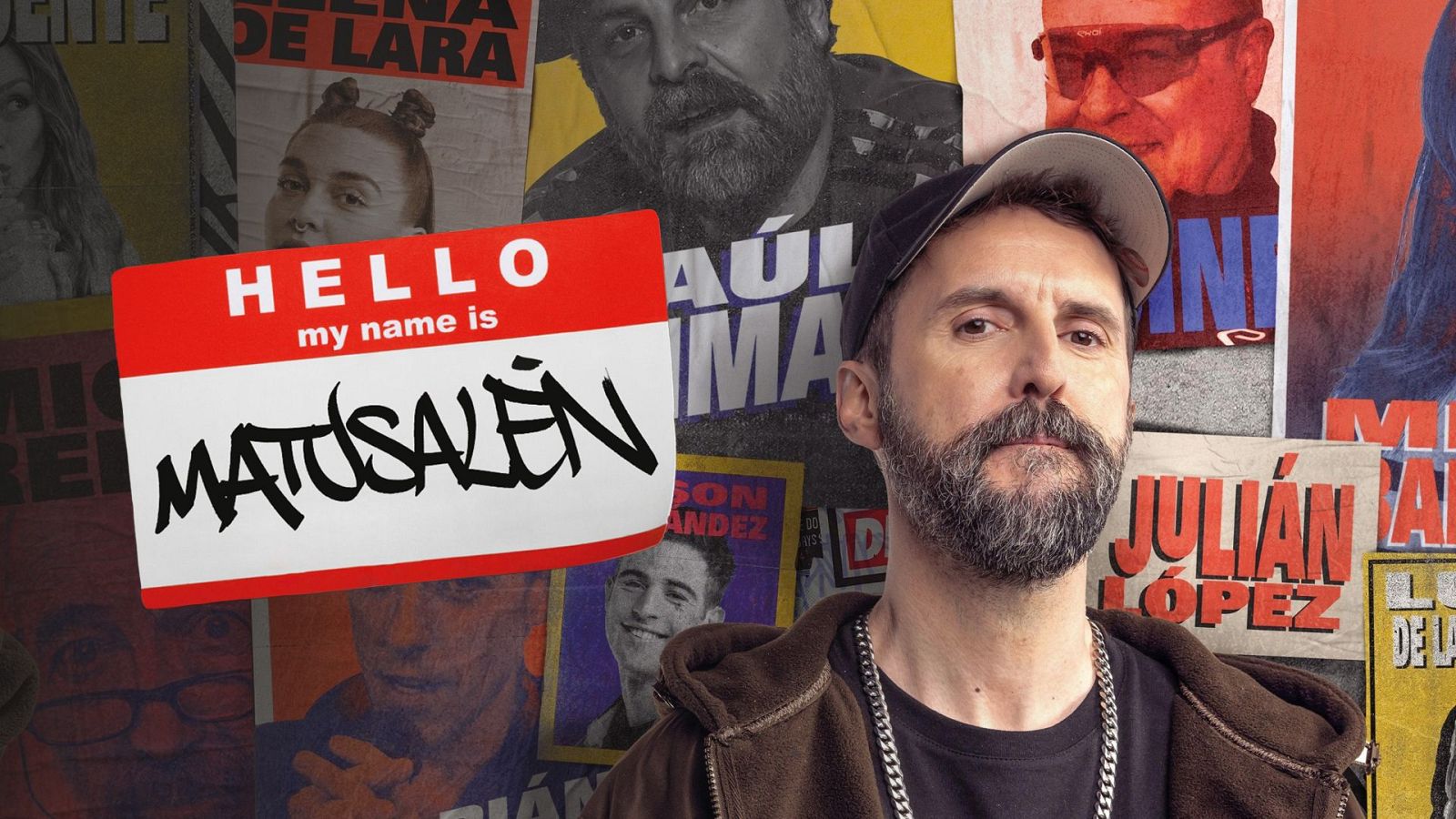 Un hombre con barba y bigote, con una gorra y chaqueta, posa junto a una etiqueta con un nombre. En el fondo, se aprecian carteles con nombres escritos.