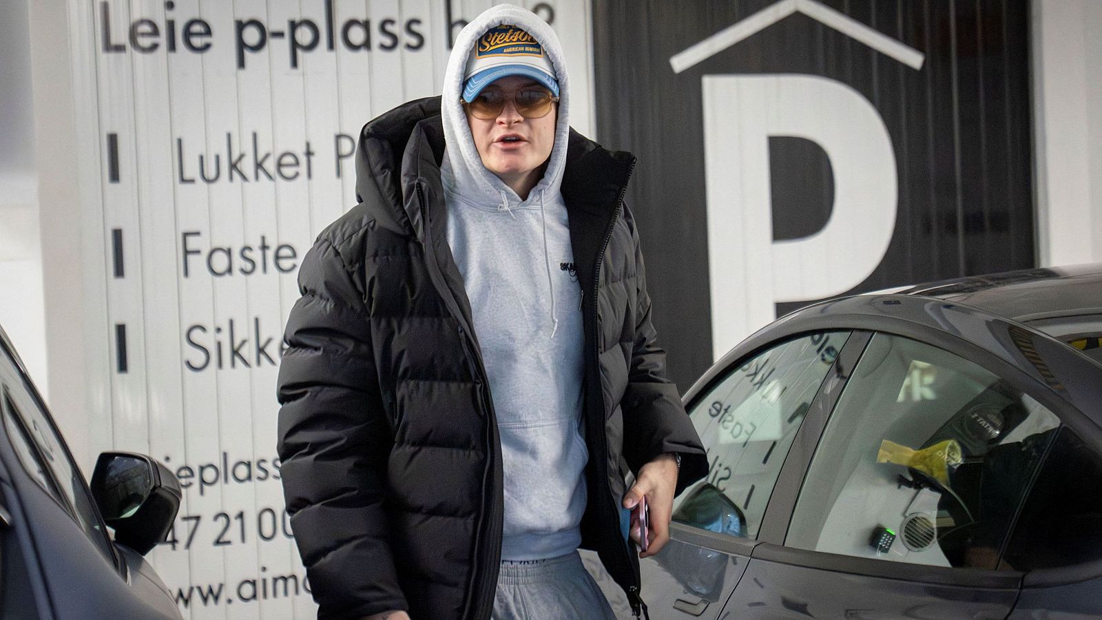 En un aparcamiento, un hombre con gafas de sol y gorra se encuentra cerca de un coche. Viste una chaqueta negra y una sudadera gris, con señales de aparcamiento y un letrero con la letra 'P' al fondo.