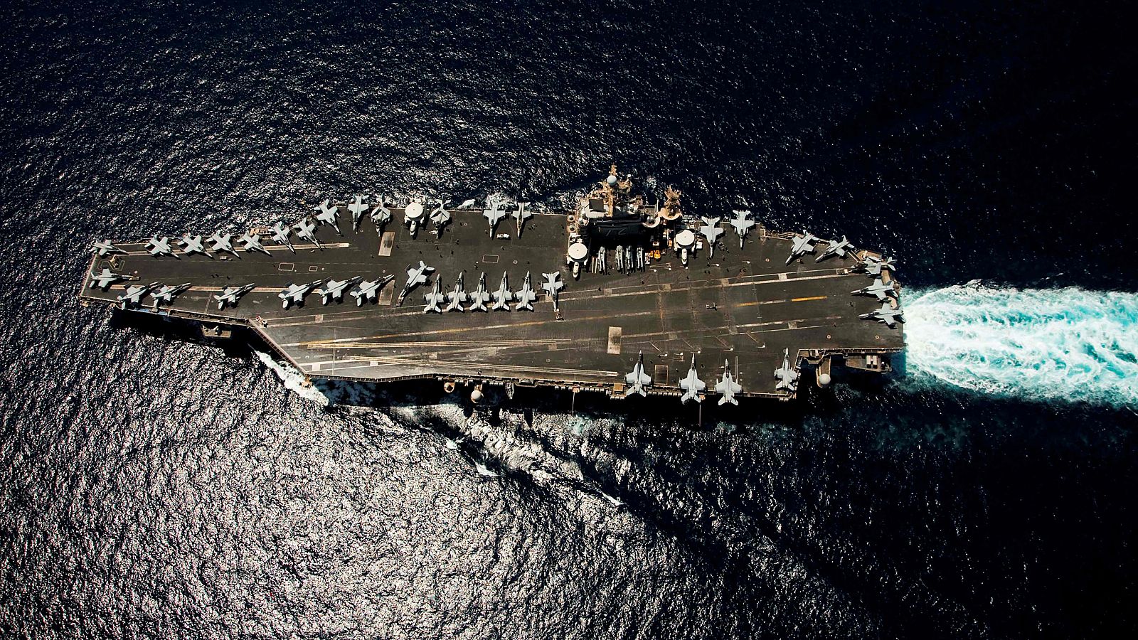 El portaaviones USS Abraham Lincoln navega por el Mar Arábigo, con aviones de combate alineados en su cubierta, en una imagen aérea que destaca el azul oscuro del mar.