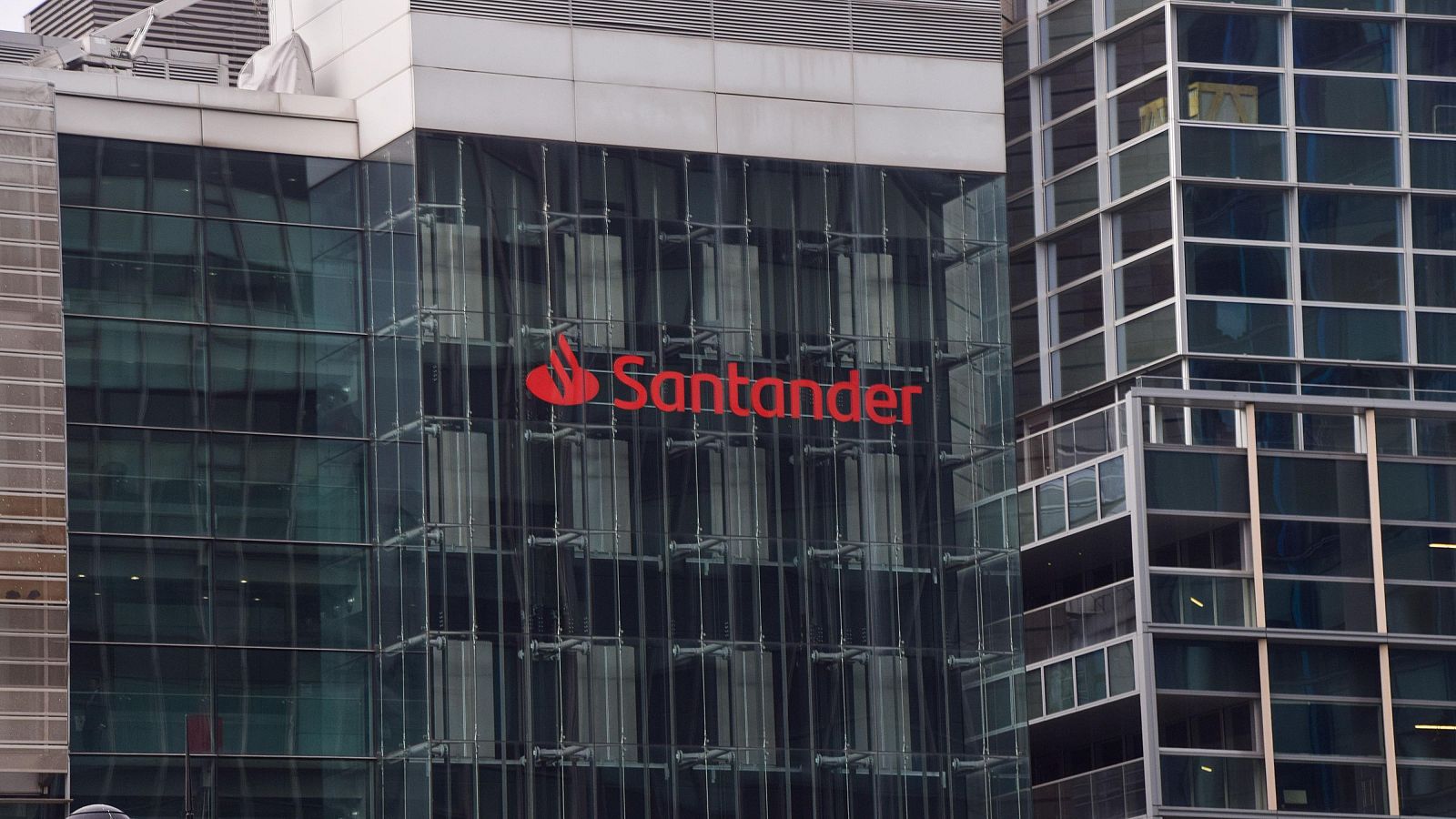 Un edificio moderno con fachada de cristal exhibe el logotipo de Santander en su parte superior, compuesto por una llama estilizada y el nombre de la entidad en letras mayúsculas.