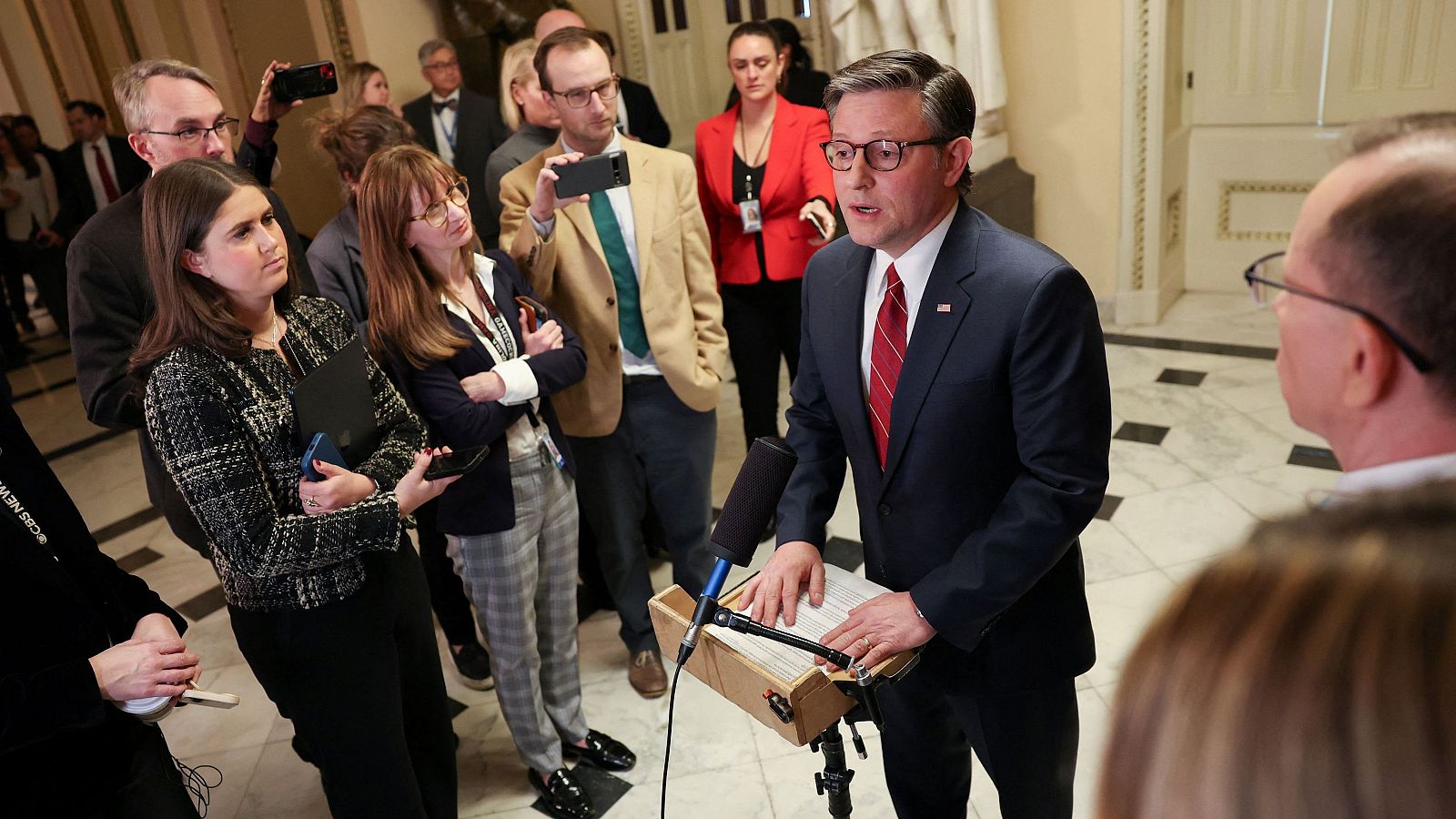 Mike Johnson, presidente de la Cámara de Representantes, habla con la prensa en el Capitolio, frente a un micrófono y un atril improvisado, rodeado de periodistas con cámaras.