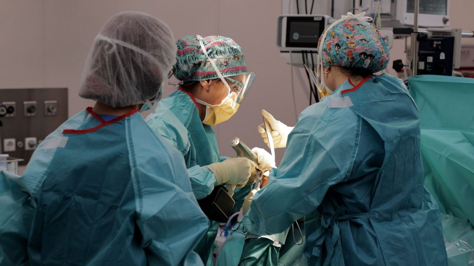 Un equip de tres professionals sanitaris, amb uniformes quirúrgics, realitzen una intervenció en un quiròfan, utilitzant instruments mèdics i amb equips de monitorització al fons.