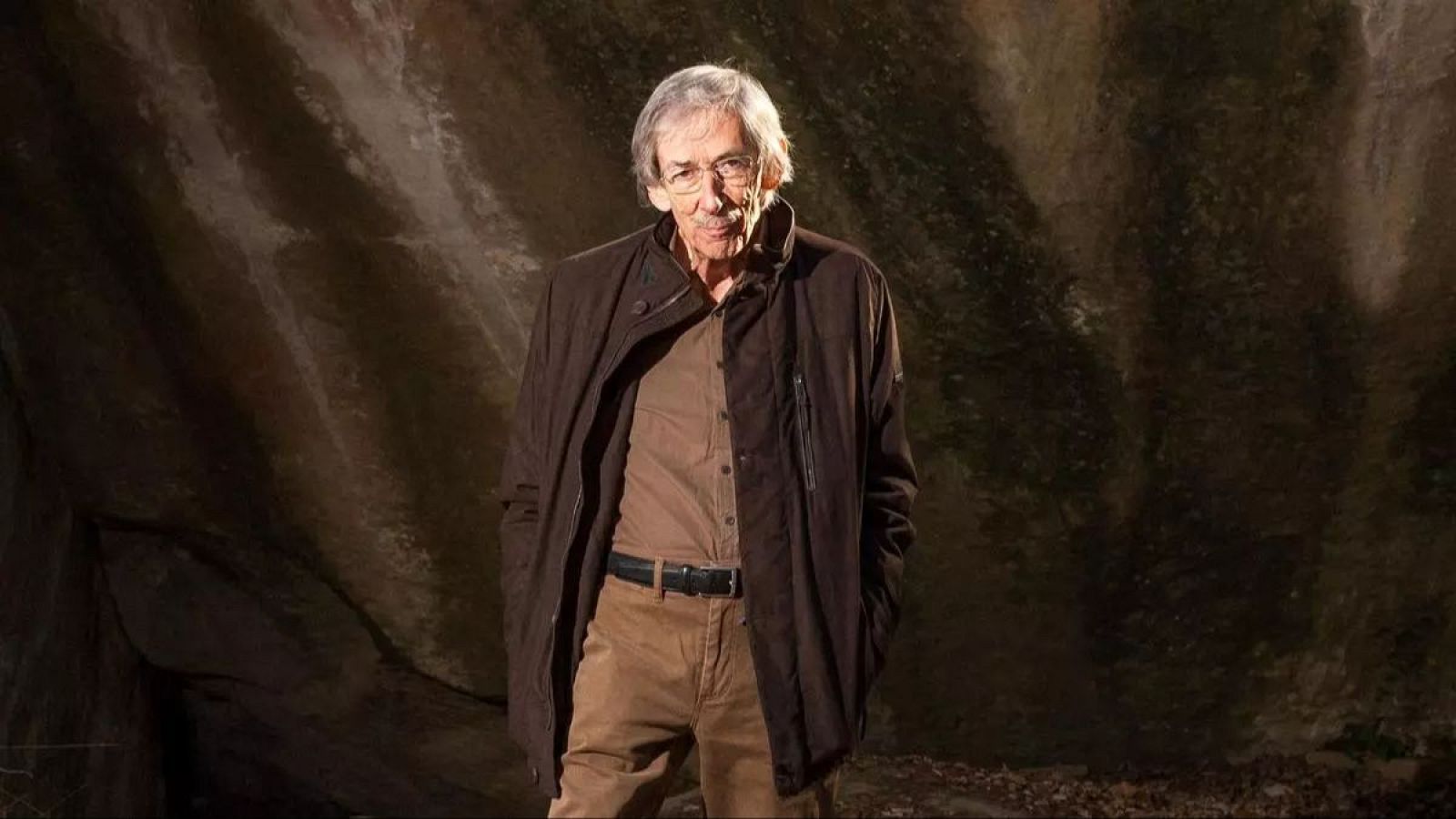 Retrato de Tomàs Pladevall, un hombre mayor con gafas y cabello canoso, vestido con camisa, pantalones y chaqueta marrones, y un cinturón negro, posando frente a una pared rocosa oscura.