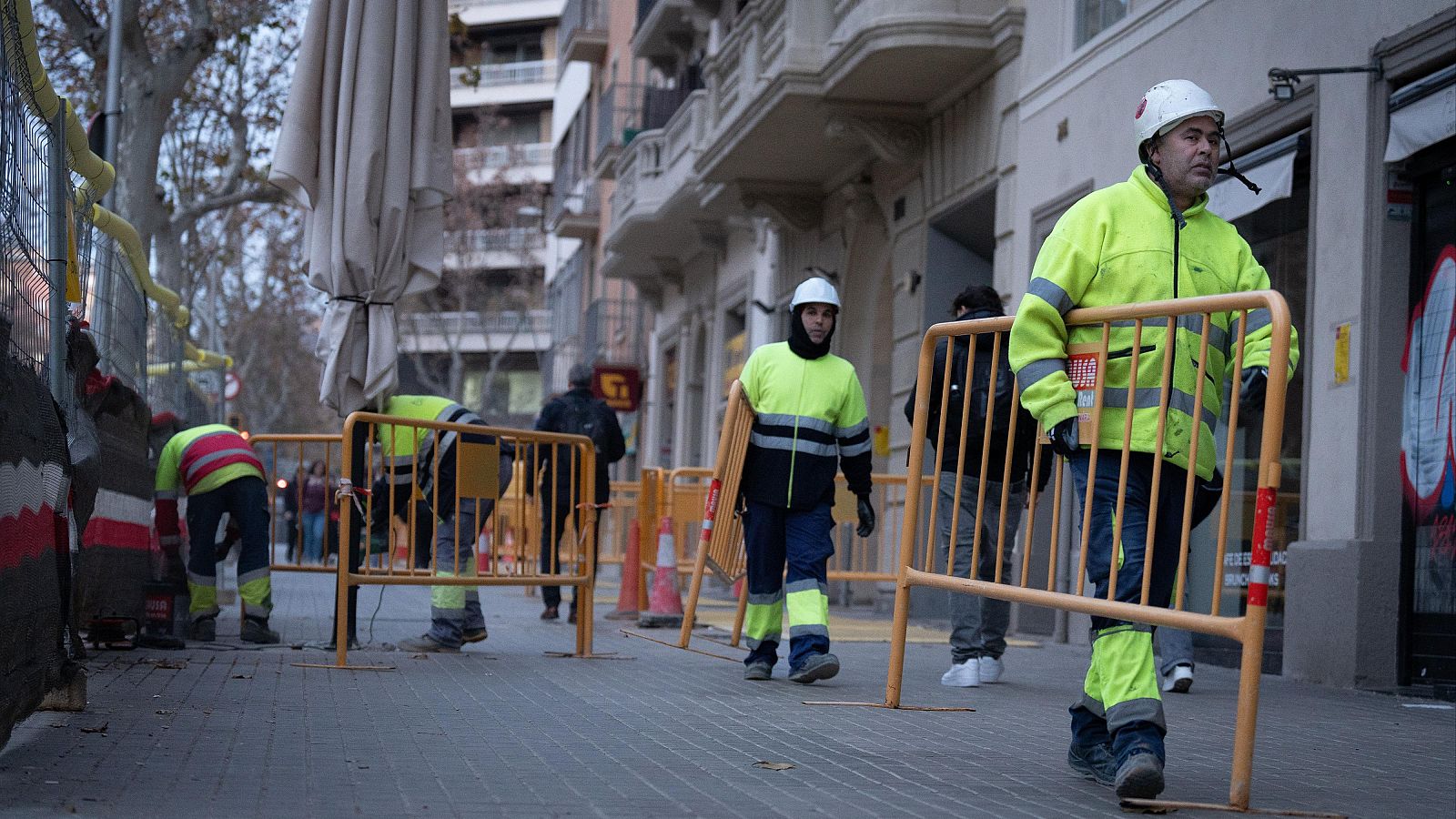 Obreros con chalecos amarillos y cascos blancos trabajan en una acera, usando vallas metálicas para delimitar una zona de obras en una calle urbana.
