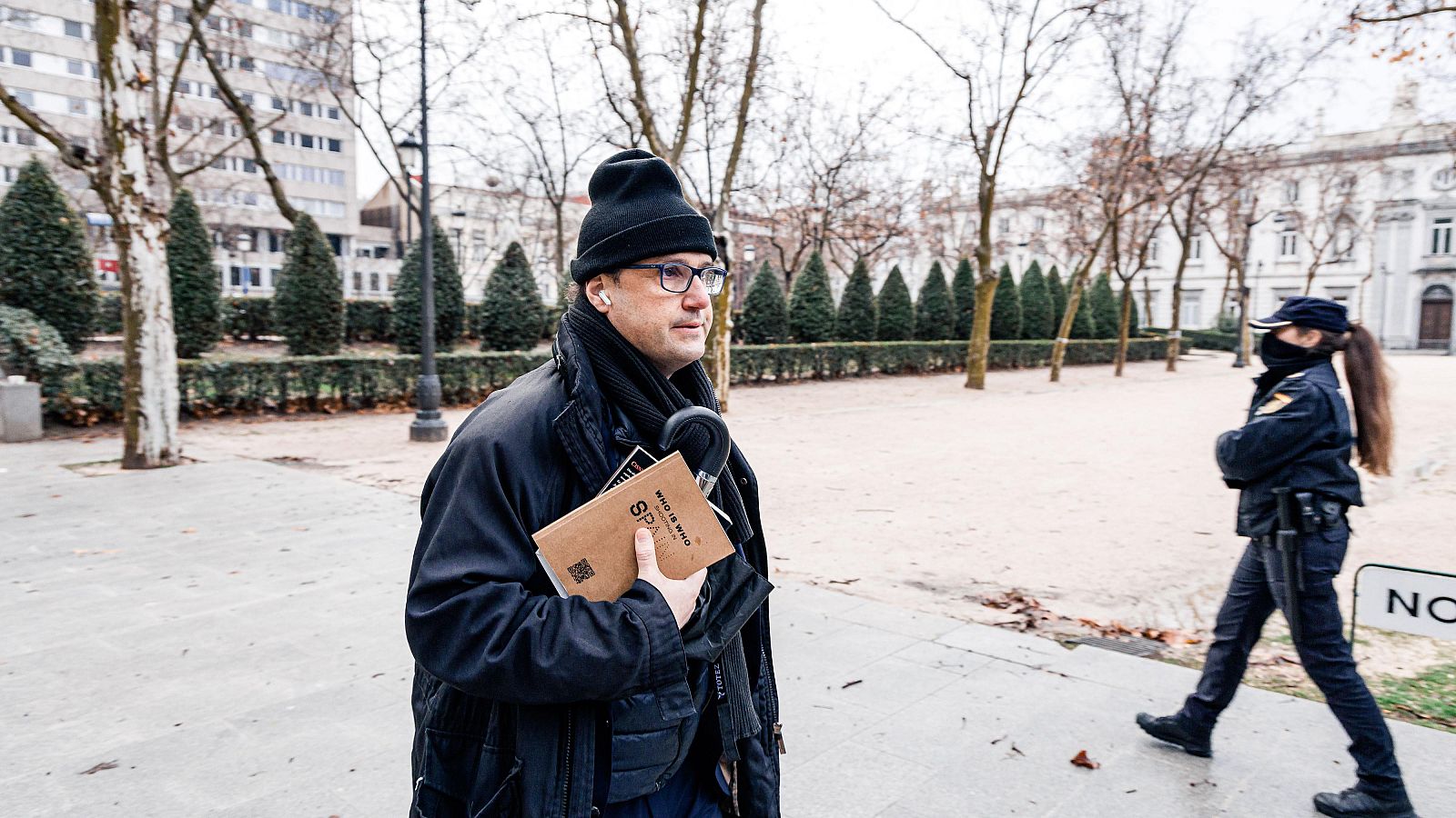 En una plaza o parque, un hombre con gafas y abrigo oscuro sostiene un libro. Una agente de policía uniformada está cerca. El ambiente es frío, con árboles sin hojas y edificios al fondo.