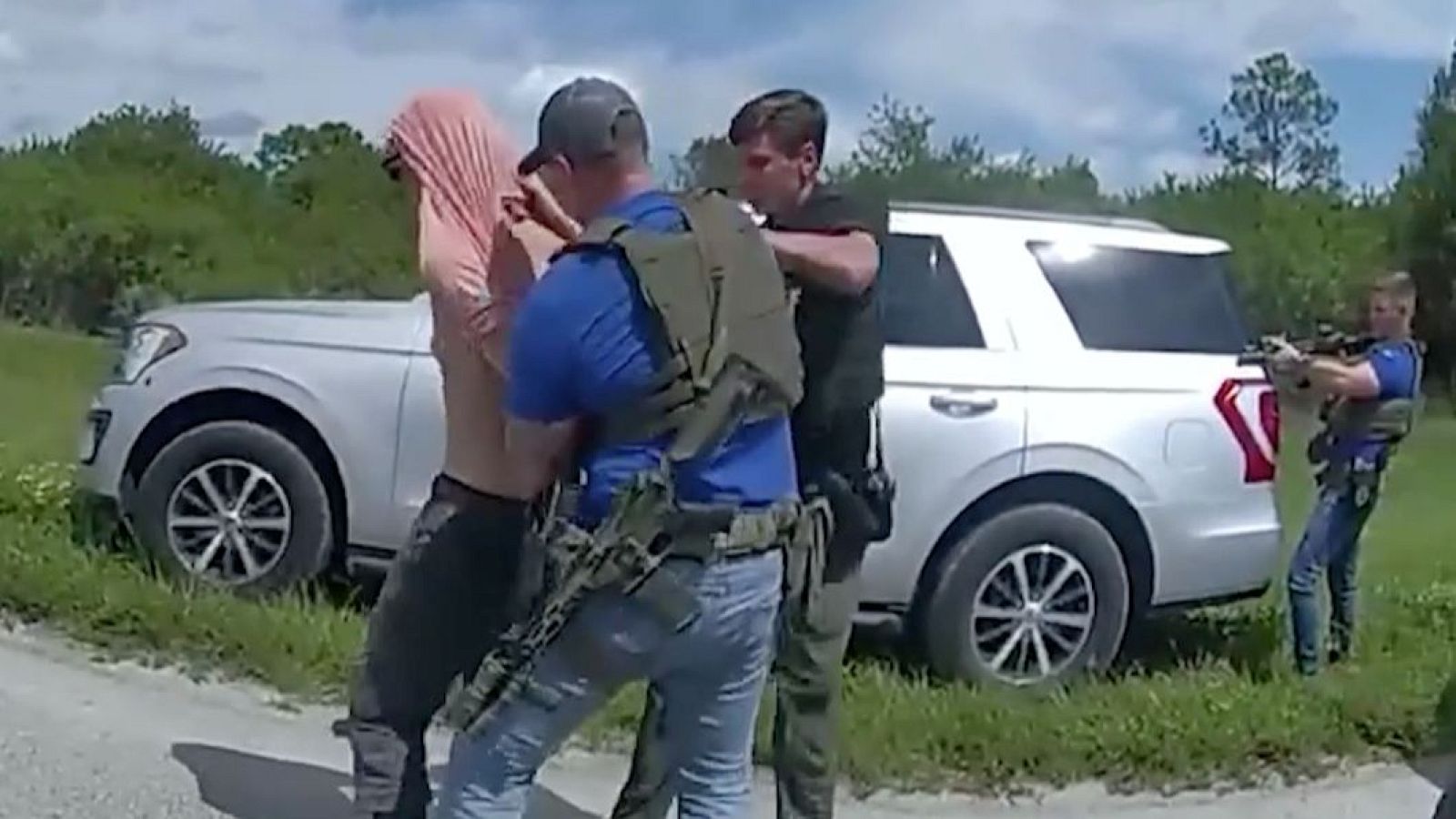 Agentes de seguridad detienen a un individuo en una zona con vegetación. El sospechoso, con una prenda rosa, es sujetado mientras un tercer agente apunta con un arma larga desde detrás de un vehículo.
