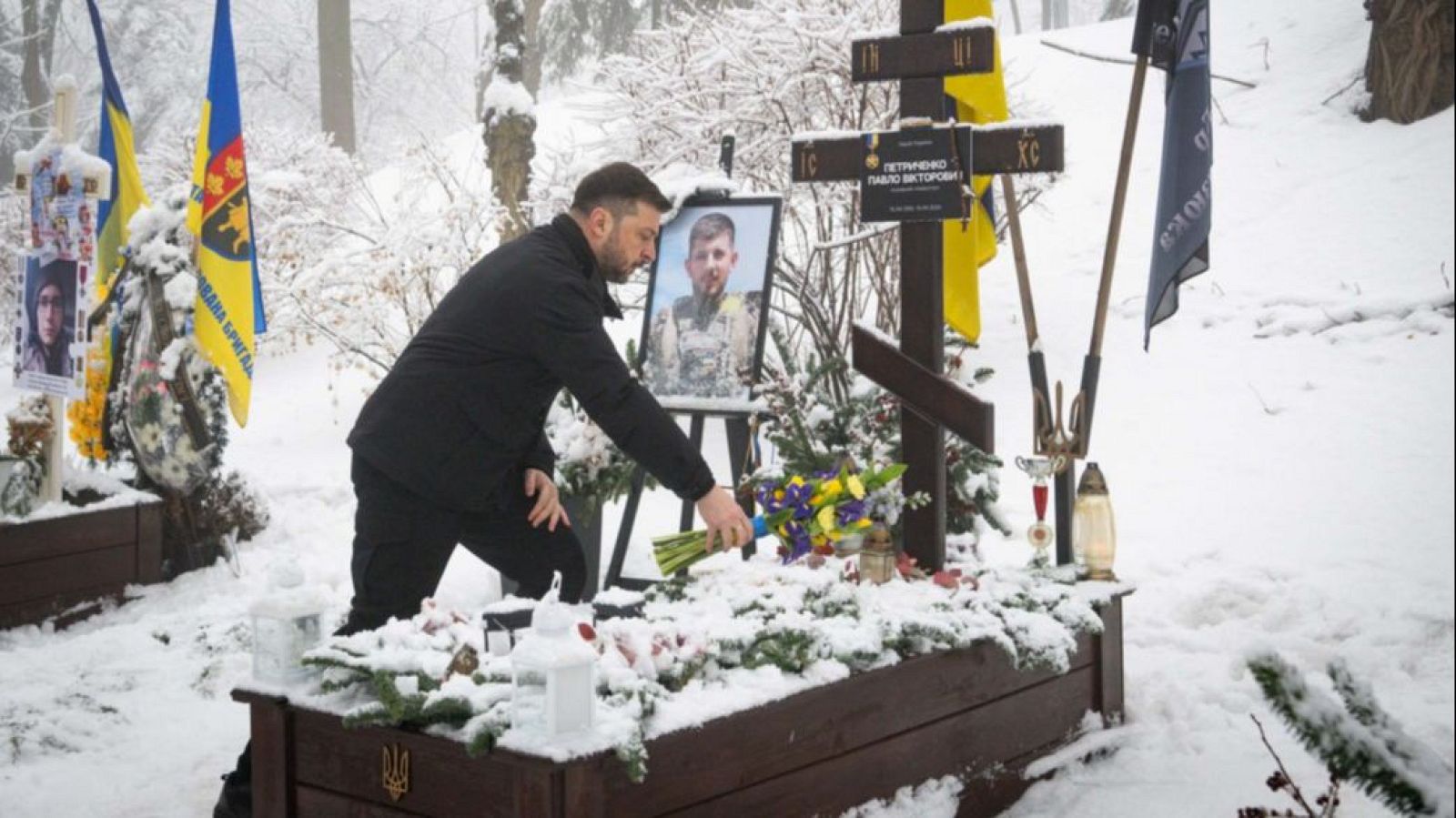 El presidente Zelenski, vestido de negro, honra la memoria de un soldado en un cementerio cubierto de nieve, depositando flores en una tumba con una cruz y una fotografía.