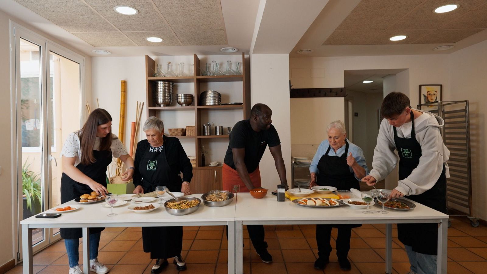 Cinc persones, vestides amb davantals, preparen menjar en una cuina lluminosa. Hi ha una taula blanca plena de plats i utensilis. Dues dones grans i dos joves, un d'ells de raça negra, estan involucrats en la preparació.