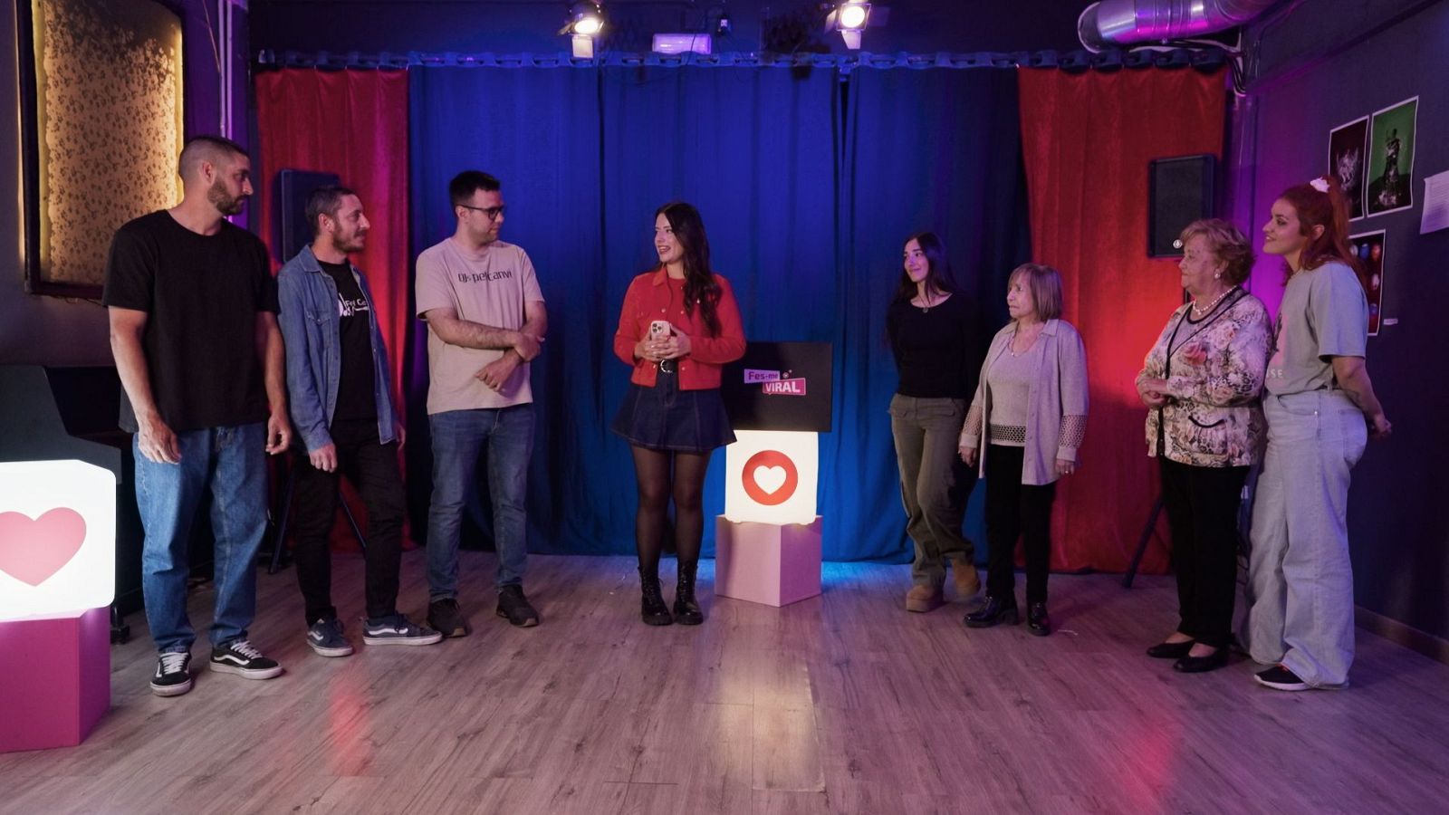 En un estudi, un grup de persones, incloent homes amb roba informal i dones amb vestits variats, semblen participar en un esdeveniment. Una dona amb jaqueta vermella sosté un telèfon, mentre que al fons hi ha decoració amb cubs i cortines.