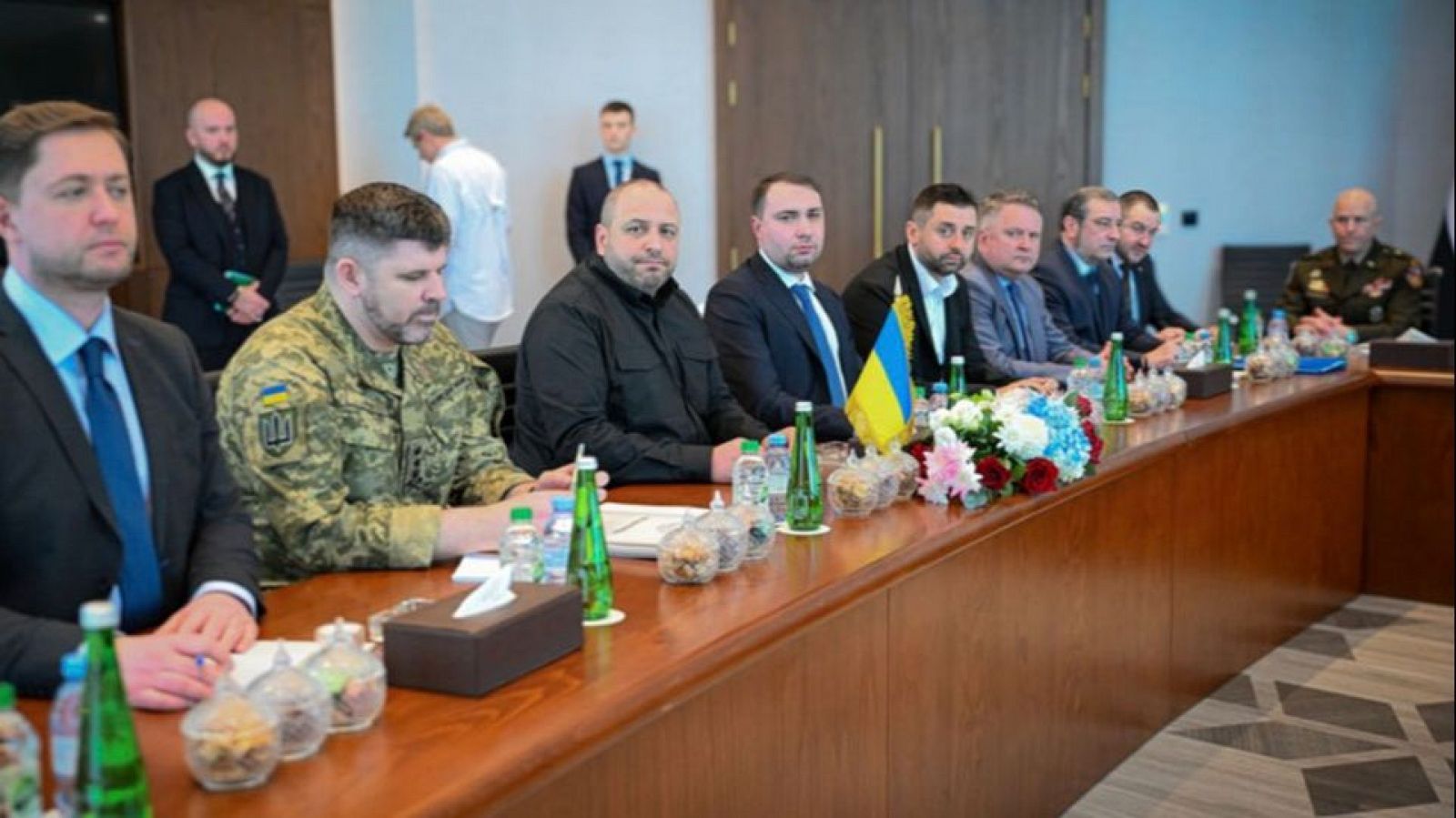 Una mesa de negociaciones en Abu Dabi es el escenario de una reunión formal entre delegados, algunos vestidos con trajes y otros con uniformes. En el centro de la mesa, se distinguen una bandera y arreglos florales.