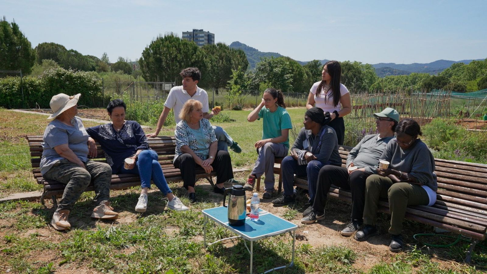 En un huerto soleado, un grupo de personas, incluyendo a mujeres con sombreros y pañuelos, y hombres con gorras, comparten un momento. Hay una mesa azul con objetos y el fondo muestra vegetación y un edificio.