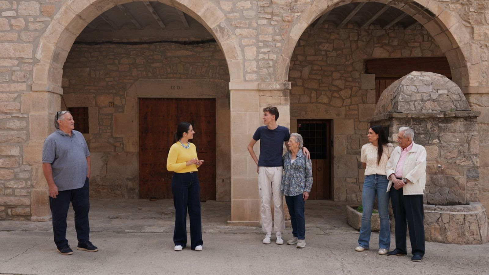 Un grup de persones posa davant d'una construcció de pedra amb arcs, amb una porta de fusta al fons. Hi ha diverses persones de diferents edats, vestides amb roba casual.