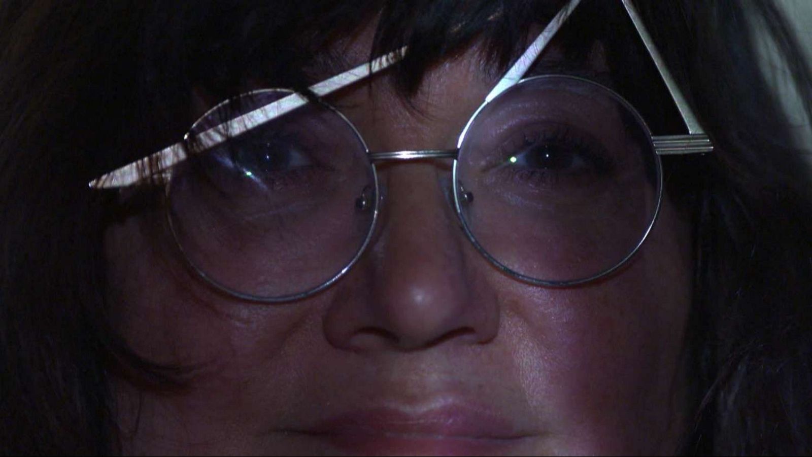 Primer plano del rostro de una mujer con gafas redondas y montura metálica, posiblemente una cineasta, con una patilla de las gafas levantada, capturado con una iluminación tenue que genera sombras.