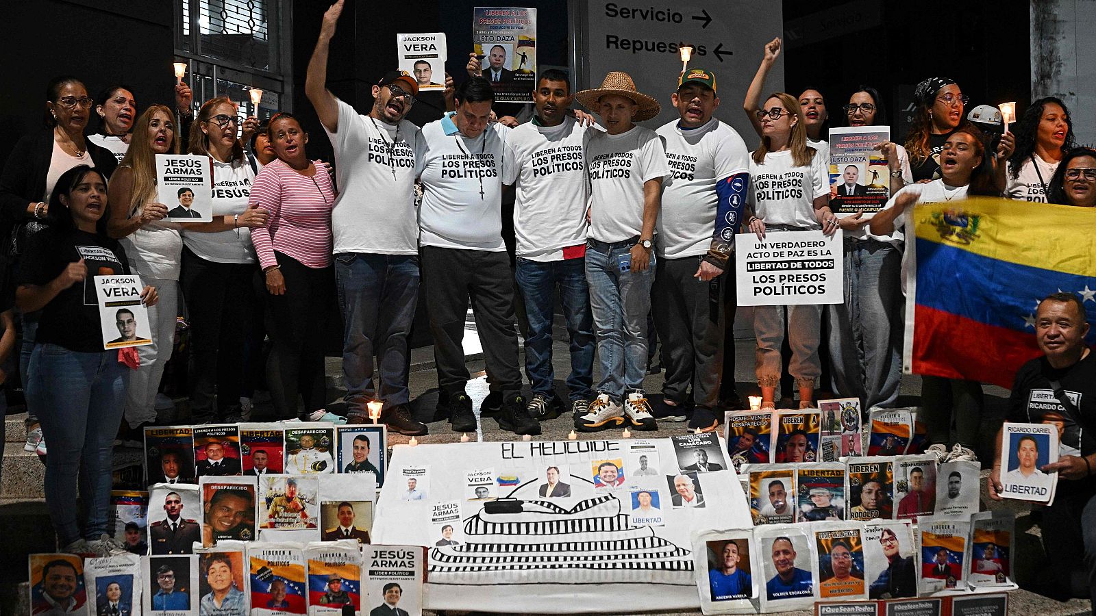 Manifestación en Caracas, frente a un edificio, con personas sosteniendo carteles y velas. Se observan fotos y mensajes relacionados con la libertad de presos políticos. Algunos manifestantes visten camisetas con consignas.