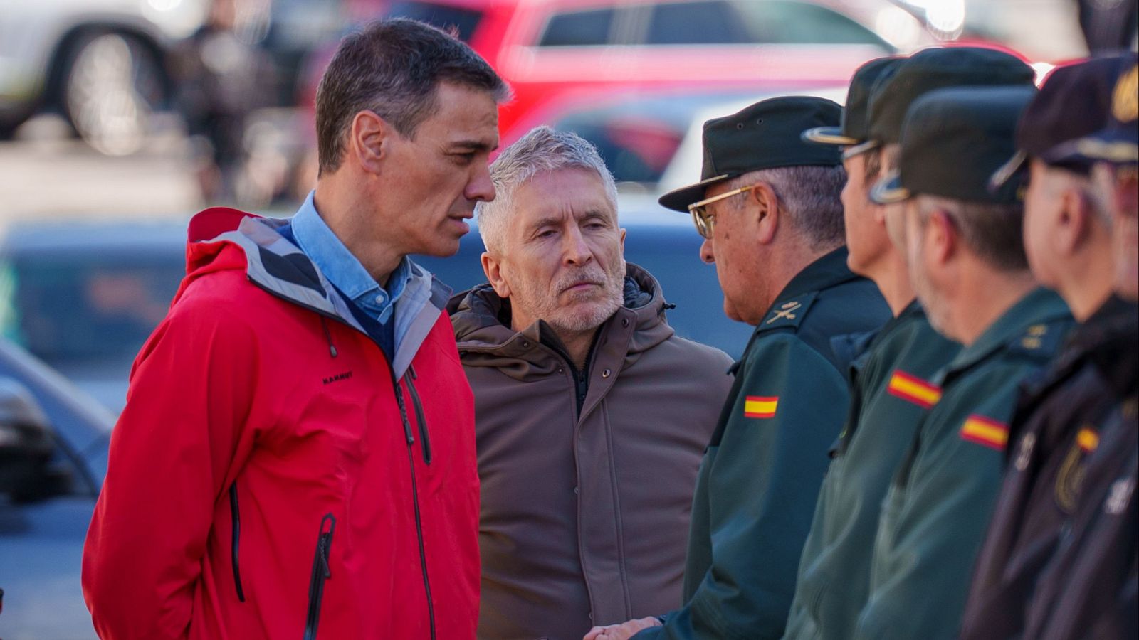 Pedro Sánchez, con chaqueta roja, saluda a agentes de seguridad uniformados en un entorno exterior. Un hombre con chaqueta marrón está a su lado, y los agentes visten uniformes verde oscuro con la bandera española.