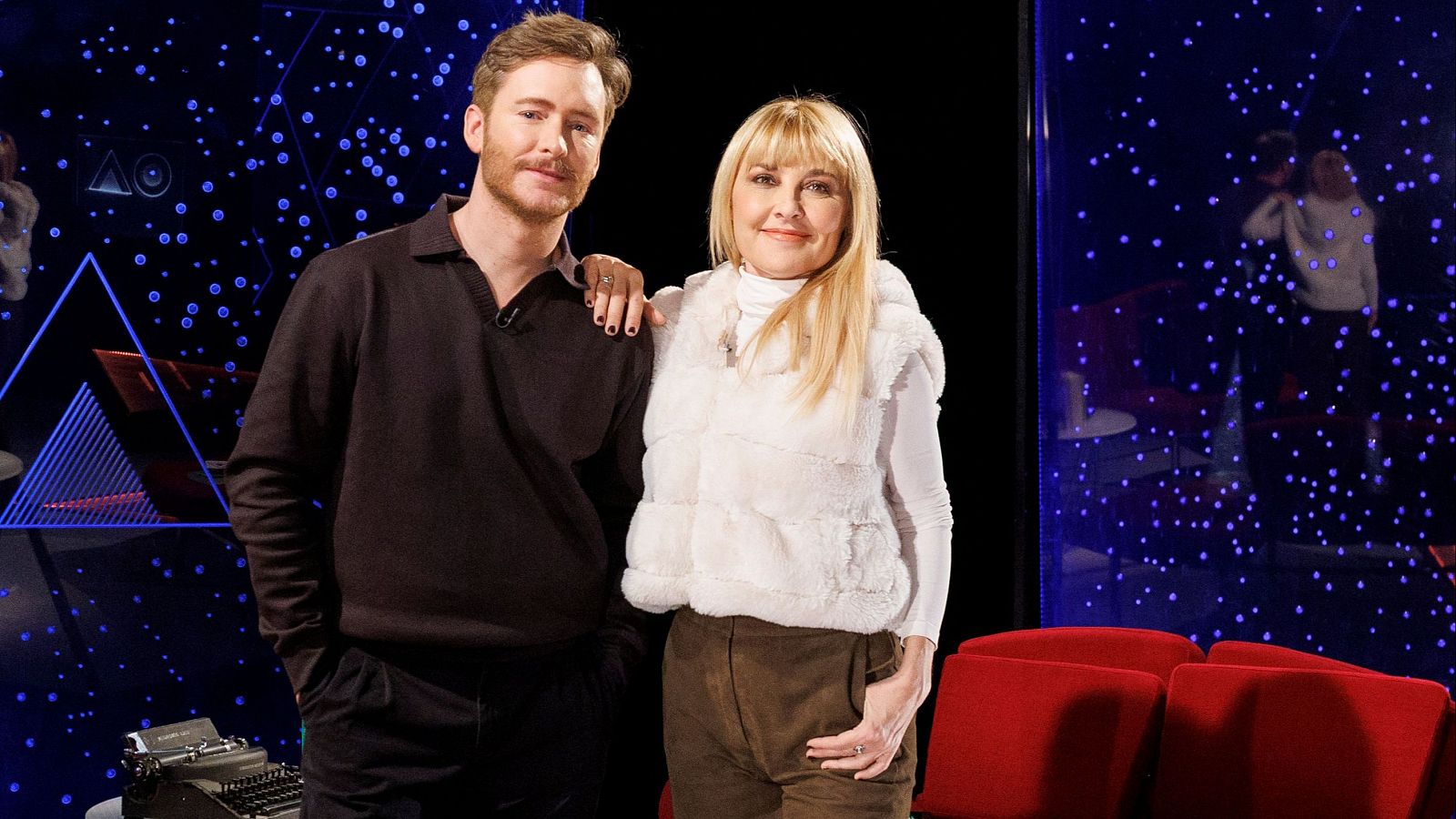 En un plató televisivo, un hombre con barba y jersey oscuro posa junto a una mujer rubia con una sonrisa. El fondo oscuro simula un cielo estrellado con luces azules y un triángulo.