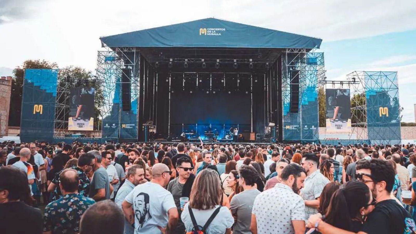 En el festival, un escenario con una lona azul y el logo del evento sirve de plataforma para una banda. Frente a él, una gran multitud observa el espectáculo, con pantallas a los lados mostrando imágenes.