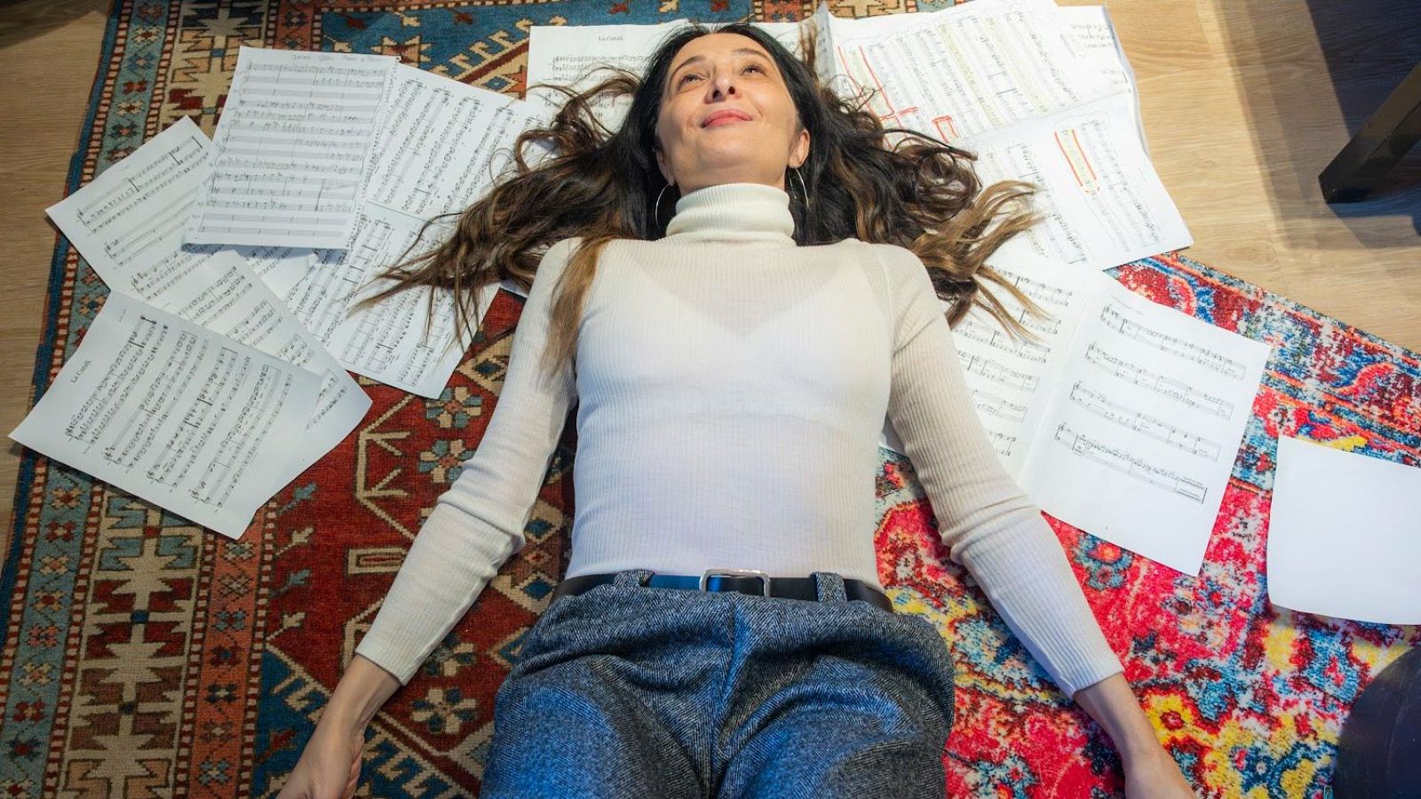 En una imatge, Giulia Valle, amb cabells castanys i vestida amb jersei blanc, descansa sobre una catifa plena de partitures, mirant cap amunt amb serenitat.