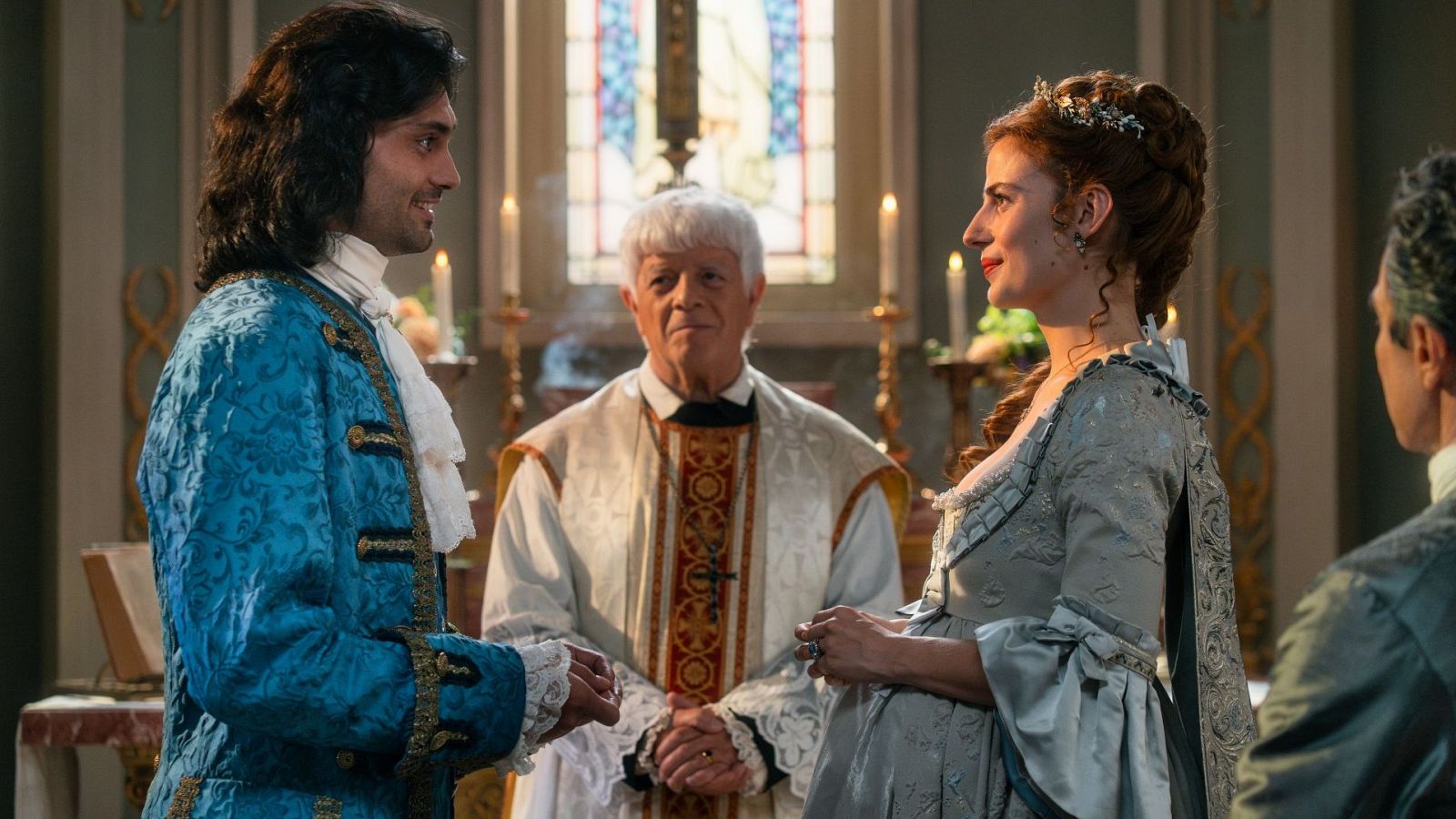En una iglesia, un sacerdote celebra una boda. Un hombre con traje azul y camisa con chorreras, y una mujer con vestido de novia grisáceo y diadema de flores, se miran sonriendo durante la ceremonia.