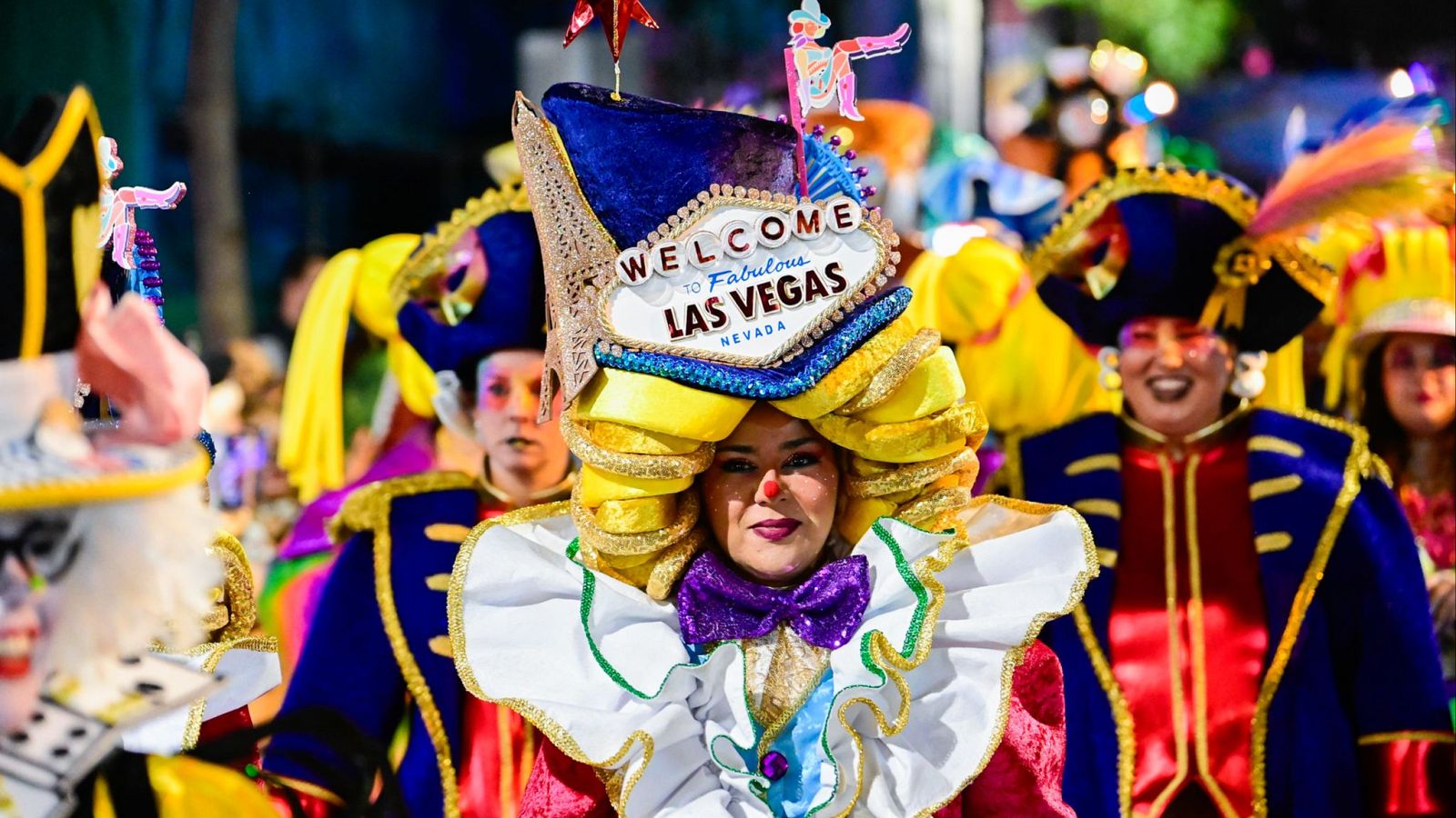 En primer plano, una mujer con maquillaje de payaso y un gran lazo morado, posa con un tocado que representa un letrero famoso. Detrás de ella, se vislumbran otros disfraces, incluyendo uno de pirata.