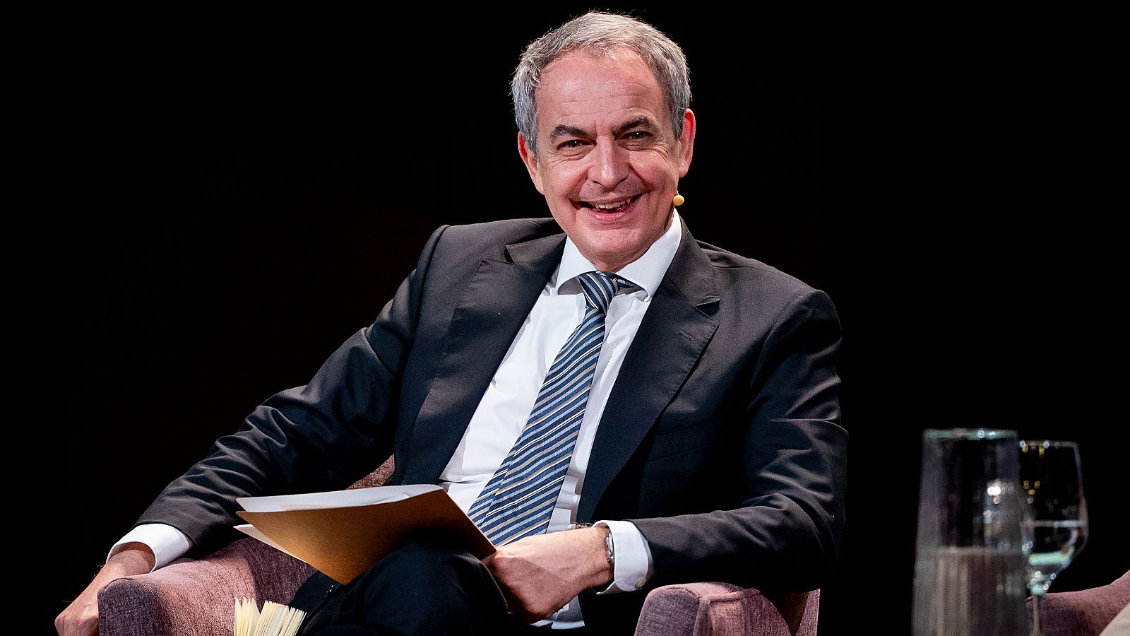 José Luis Rodríguez Zapatero, sonríe mientras está sentado en una butaca, sosteniendo un libro y un documento. Viste traje oscuro, camisa blanca y corbata a rayas, con dos vasos a su lado.