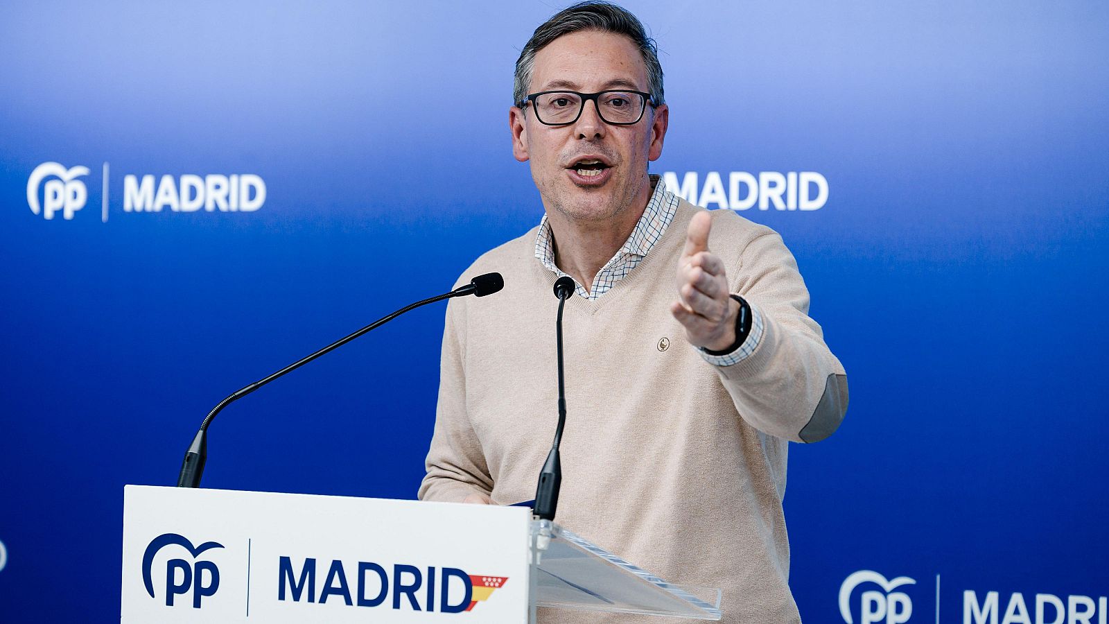 Alfonso Serrano, con gafas y jersey crema, habla desde un atril con el logo del PP de Madrid, con un fondo azul que también muestra el logo del partido y la palabra "Madrid".