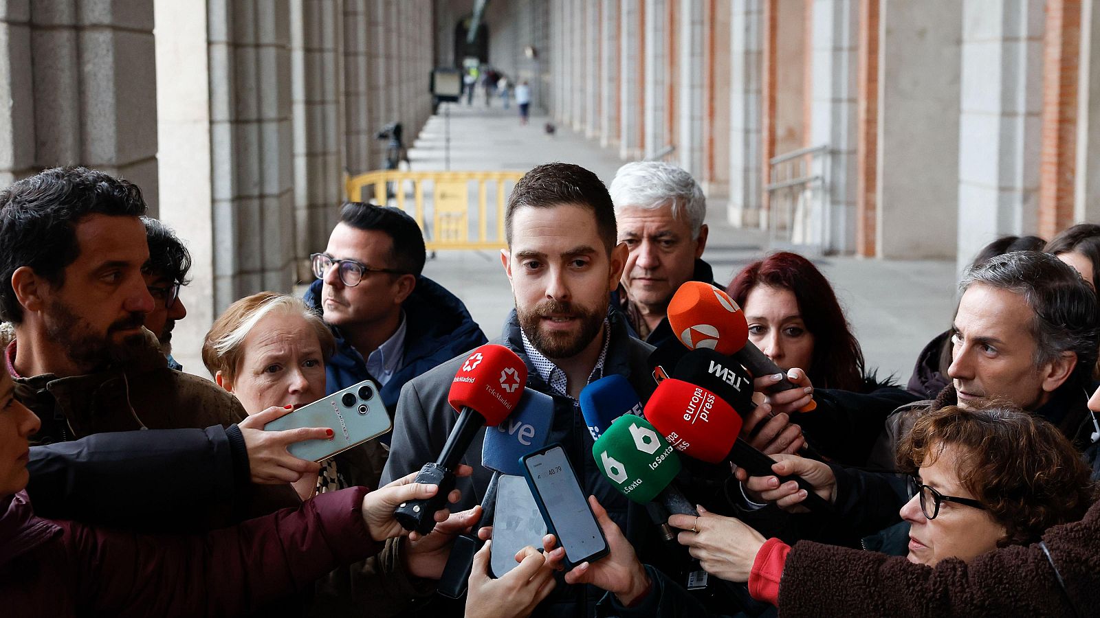 Un hombre con barba y chaqueta gris se encuentra rodeado de micrófonos de diversos medios, como Telemadrid, TVE, La Sexta y Onda Cero, mientras parece estar hablando. Varias personas sostienen micrófonos y teléfonos móviles, grabando la escena.