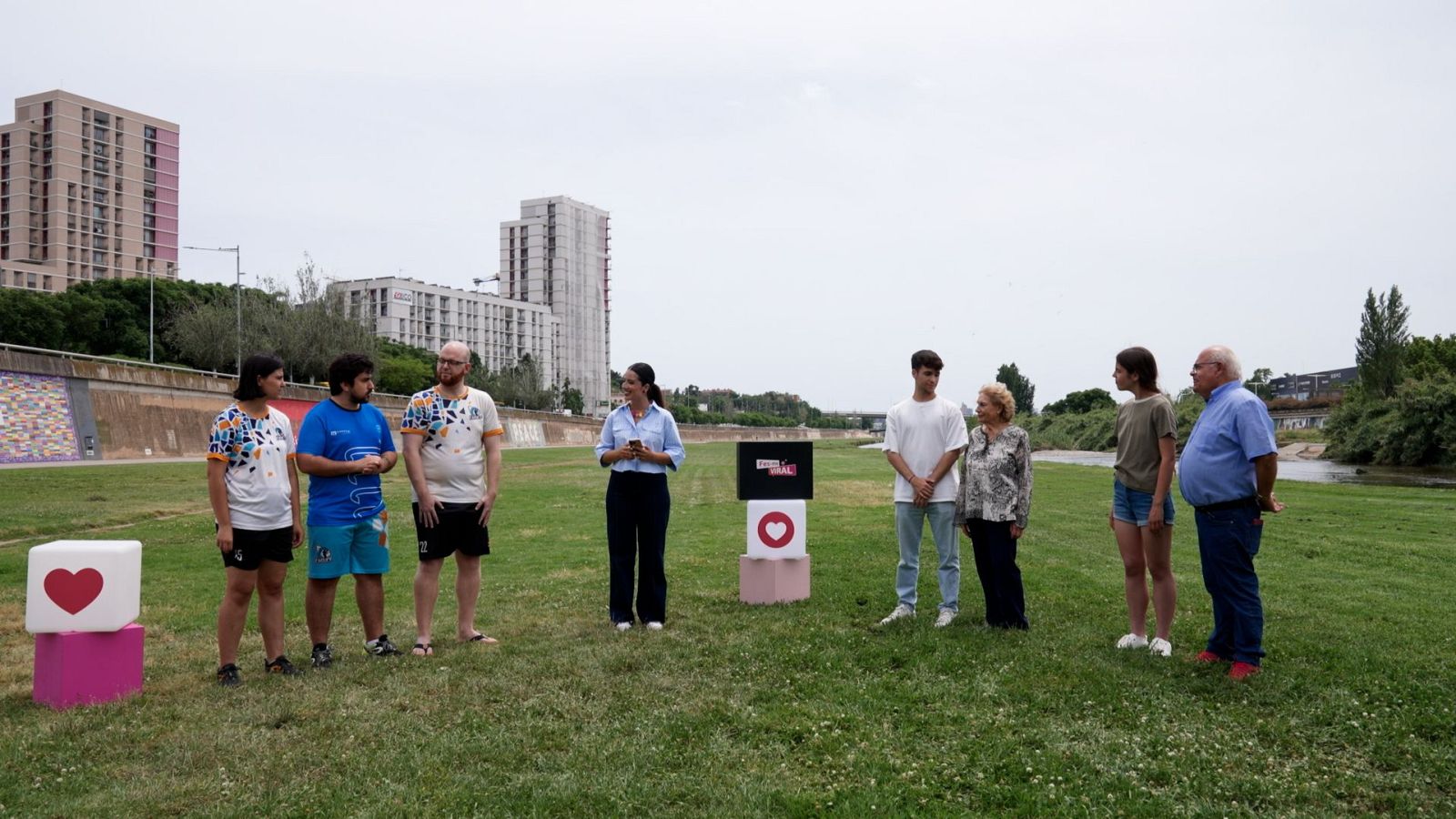 Un grup de persones es reuneixen en un camp, possiblement per a un esdeveniment. Hi ha persones de diverses edats, algunes mirant els seus telèfons, amb un suport que diu "Fes-me viral".