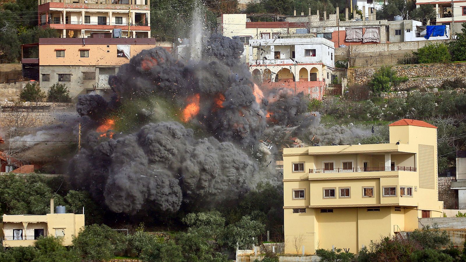 Una fuerte explosión en Ain Qana, Líbano, produce una densa columna de humo y fuego, afectando a edificios cercanos y vegetación. La imagen muestra el resultado de un evento destructivo en una zona residencial.