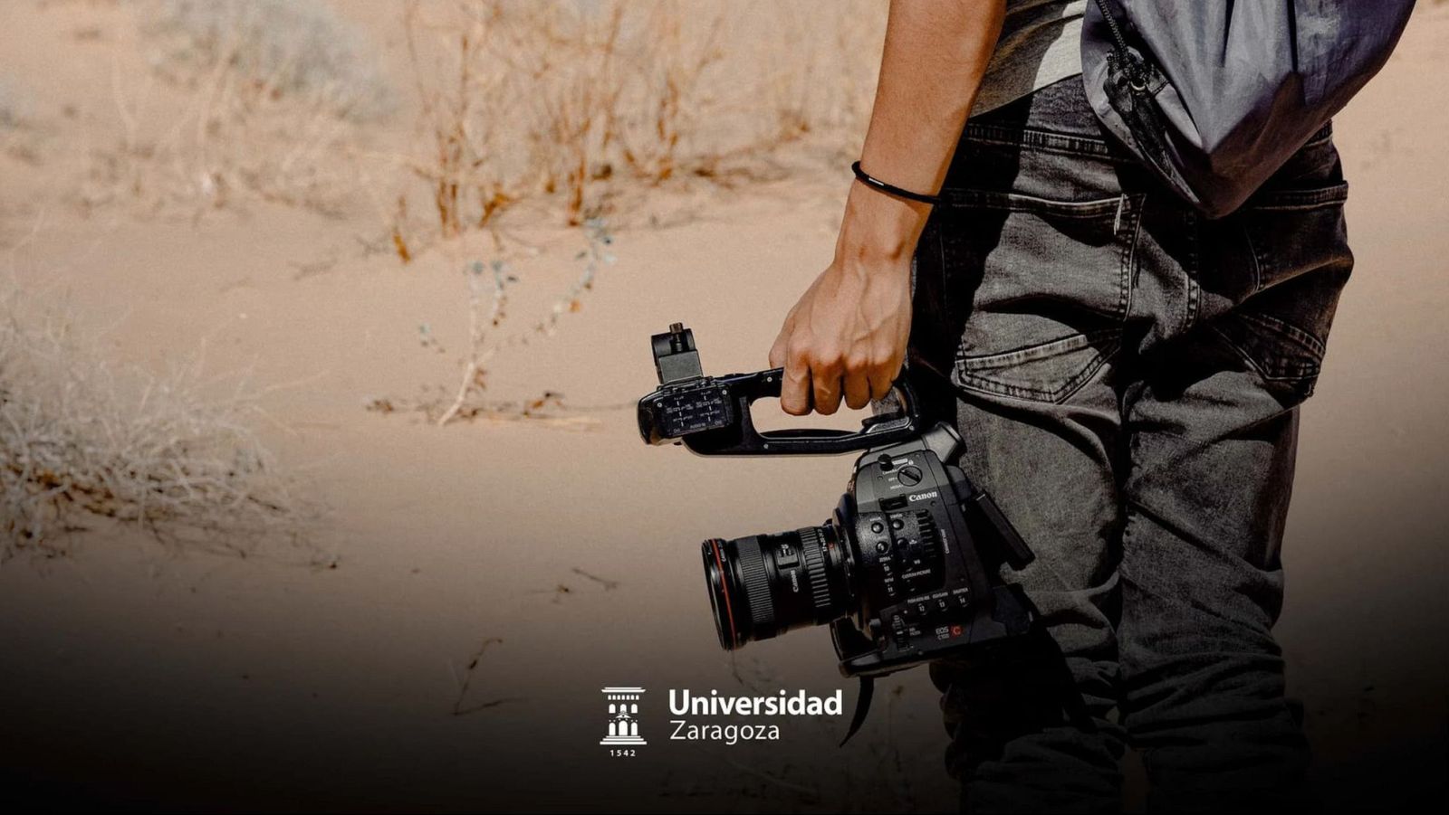 Una persona con una cámara de video Canon en un entorno desértico, con una mochila y una pulsera negra. En la imagen también se observa el logo de una universidad y la palabra "Másteres".