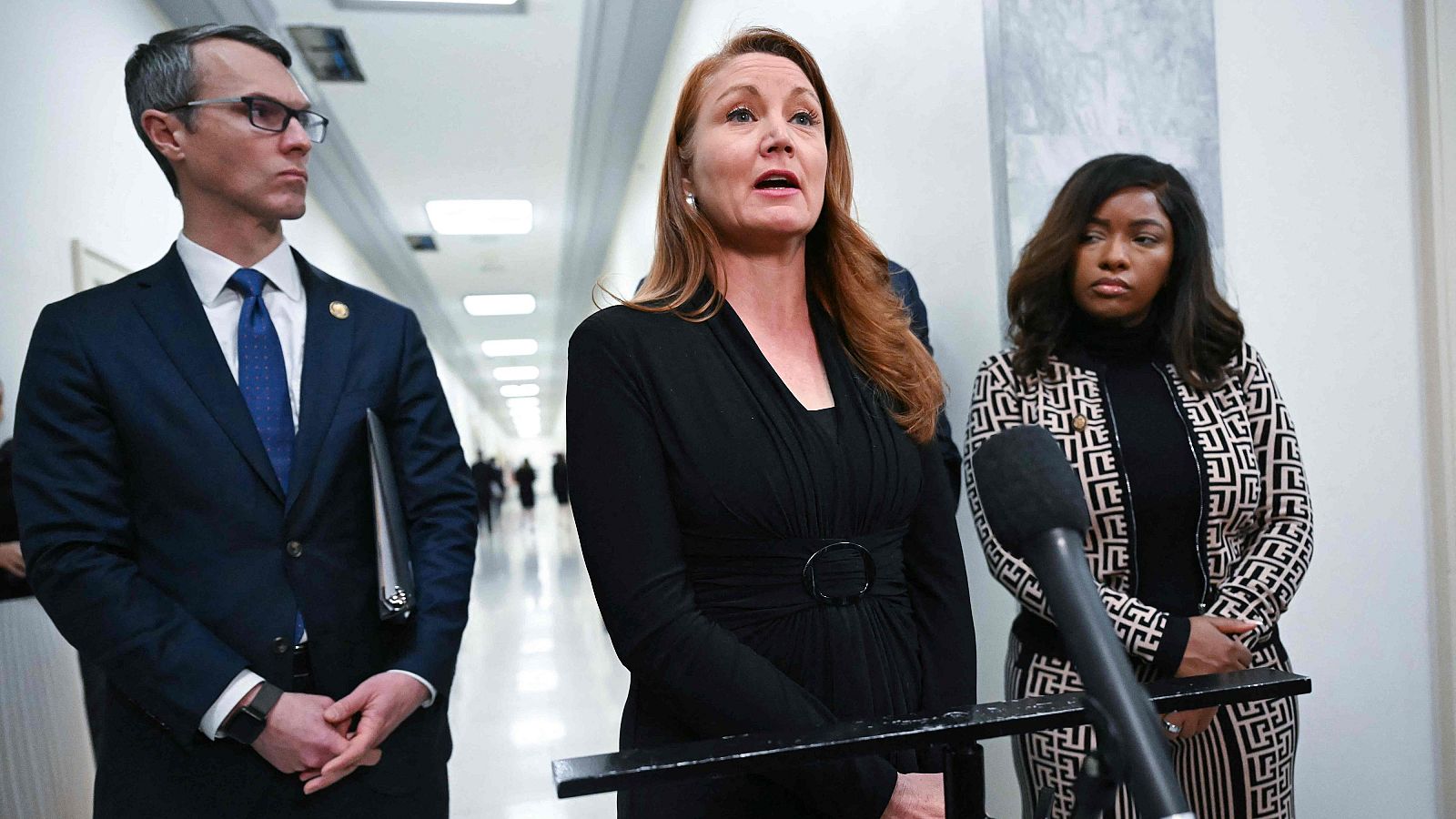 En un pasillo, una mujer pelirroja con vestido negro y micrófono está acompañada por un hombre con traje y carpeta, y otra mujer con chaqueta estampada.