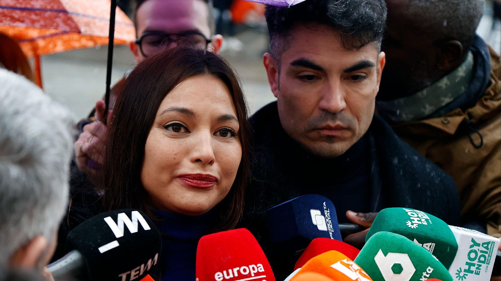 Elisa Mouliaá, rodeada de micrófonos, es abordada por la prensa en el exterior de un edificio. La escena, con fotógrafos y paraguas, sugiere un día lluvioso mientras la actriz parece estar respondiendo a las preguntas.