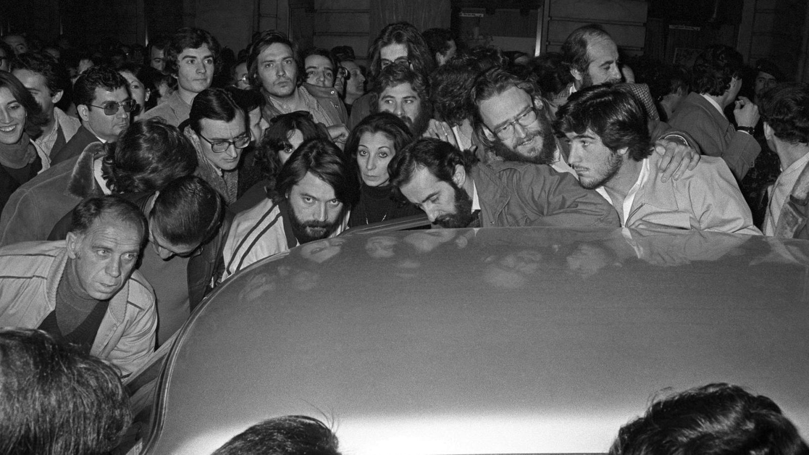 Un numeroso grupo de personas, con vestimenta variada, rodea un coche claro, prestando atención a algo que emana de él. Se observan hombres y mujeres de diferentes edades, algunos con gafas y barba, en una escena de aparente expectación.