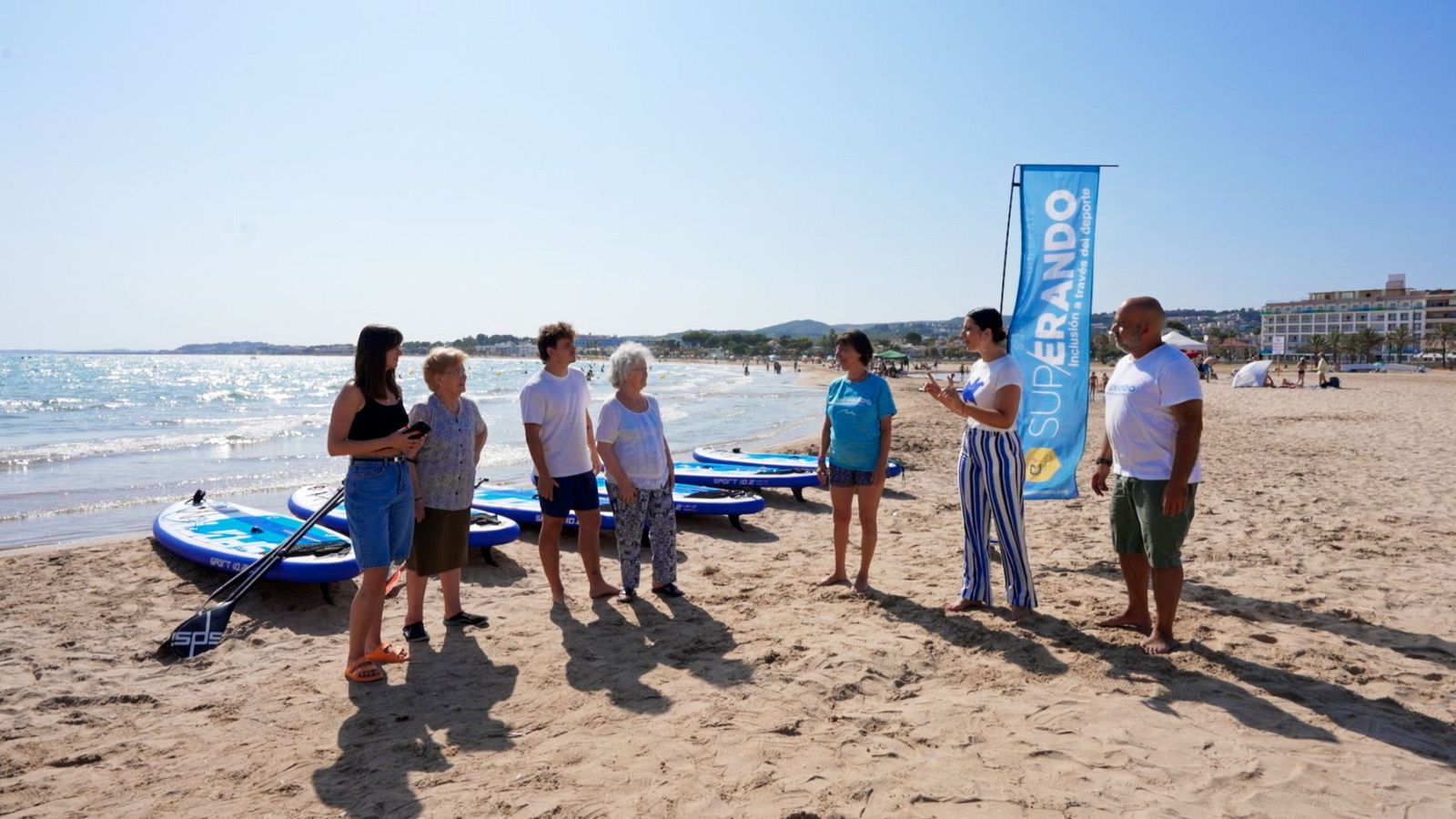 Un grup de persones, incloent-hi concursants d'un programa, es troben a la platja a prop de taules de paddle surf, amb el mar i una bandera al fons.