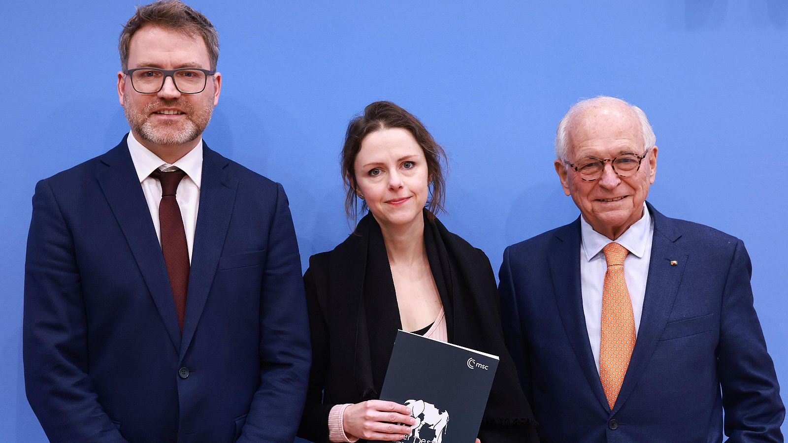 Tres personas, dos hombres con trajes y gafas y una mujer con abrigo negro, posan frente a un fondo azul. La mujer sostiene un documento con un logo.