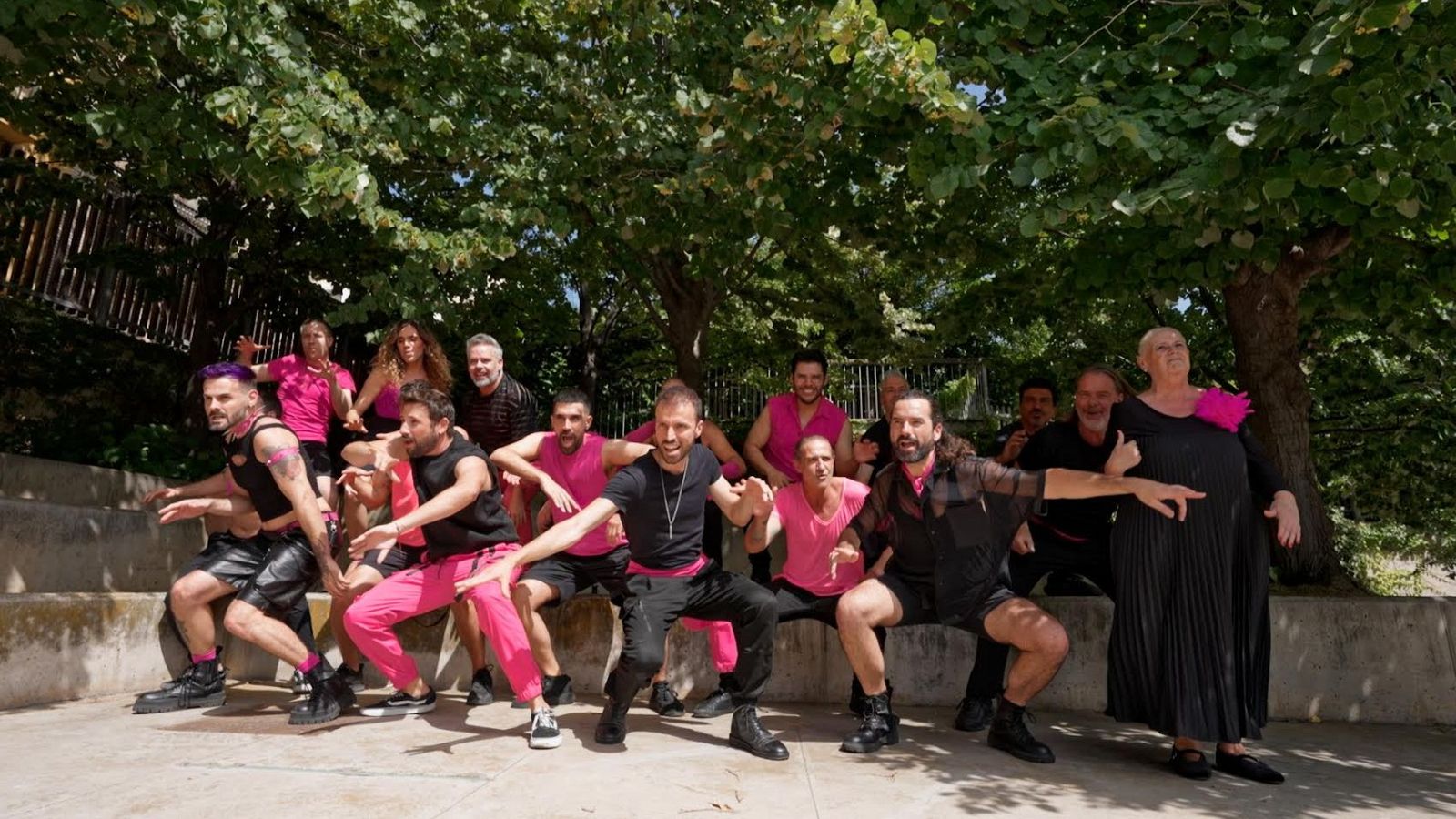 El Barcelona Gay Men's Chorus posa en un anfiteatro al aire libre. Algunos miembros visten de rosa y negro, mientras que otros visten completamente de negro, posiblemente durante una sesión de fotos o grabación.