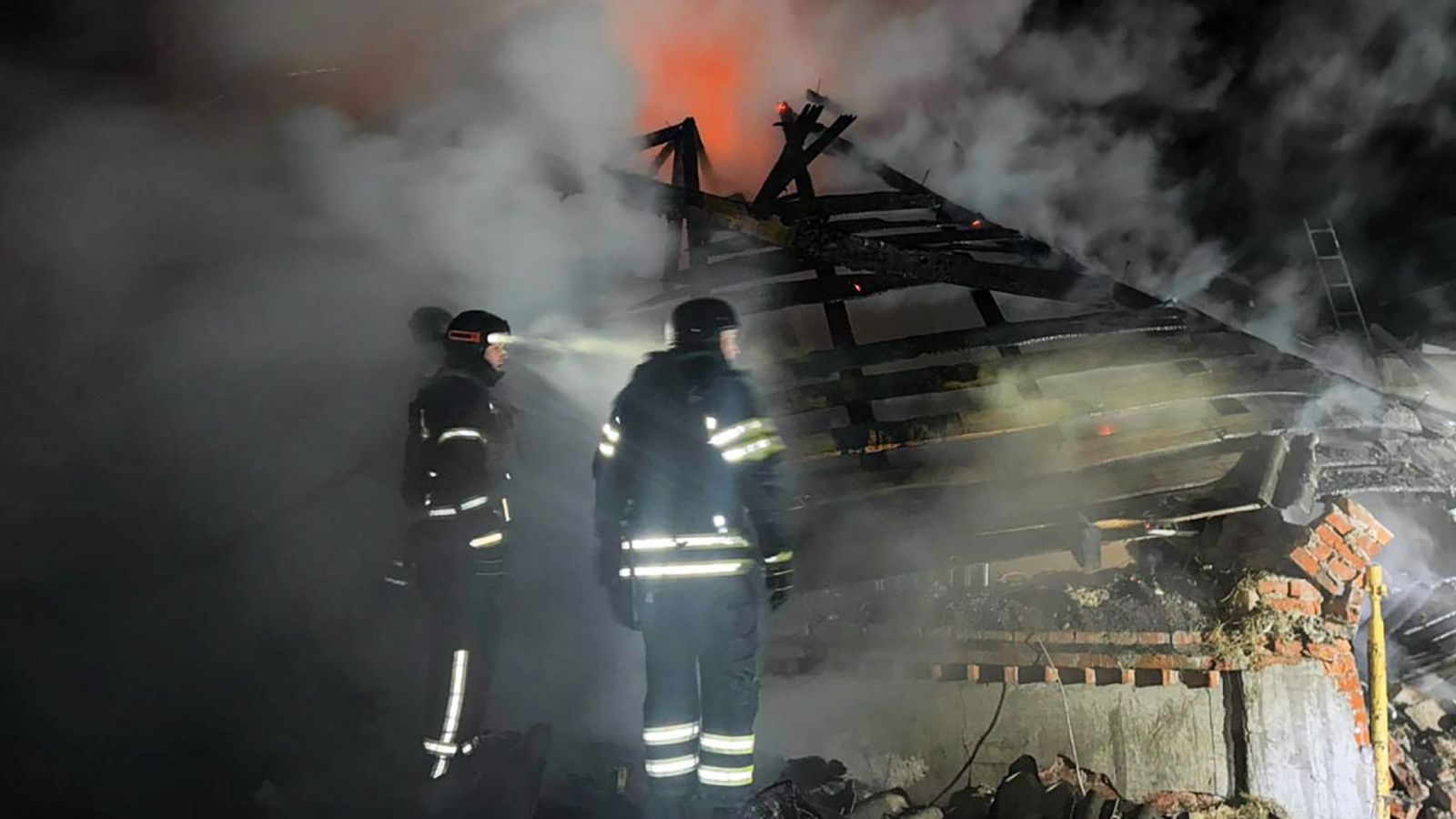 En la oscuridad de la noche, dos bomberos luchan contra un incendio en un edificio dañado, con humo denso y escombros visibles en la escena.