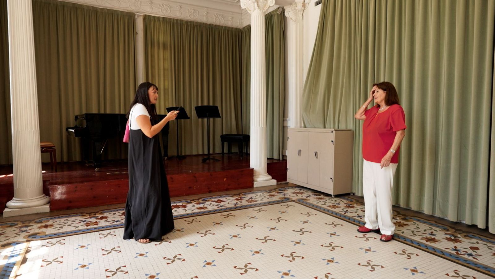 En un saló clàssic, dues dones comparteixen espai: una jove amb vestit negre i una dona major amb roba vermella i blanca. Un escenari amb un piano de cua i cortines verdes completa l'escena.